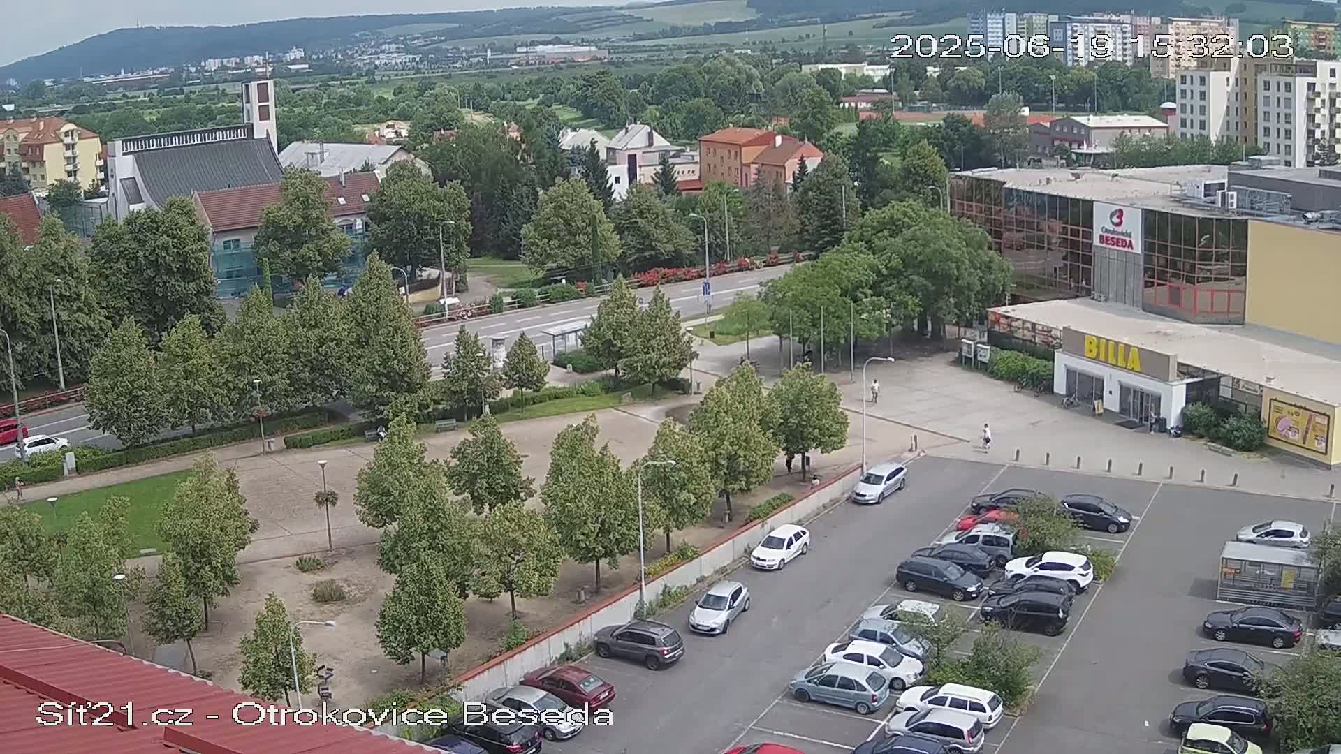 An aerial view shows a town square with a parking lot filled with cars, several buildings including a church and a supermarket, and many green trees under a partly cloudy sky.