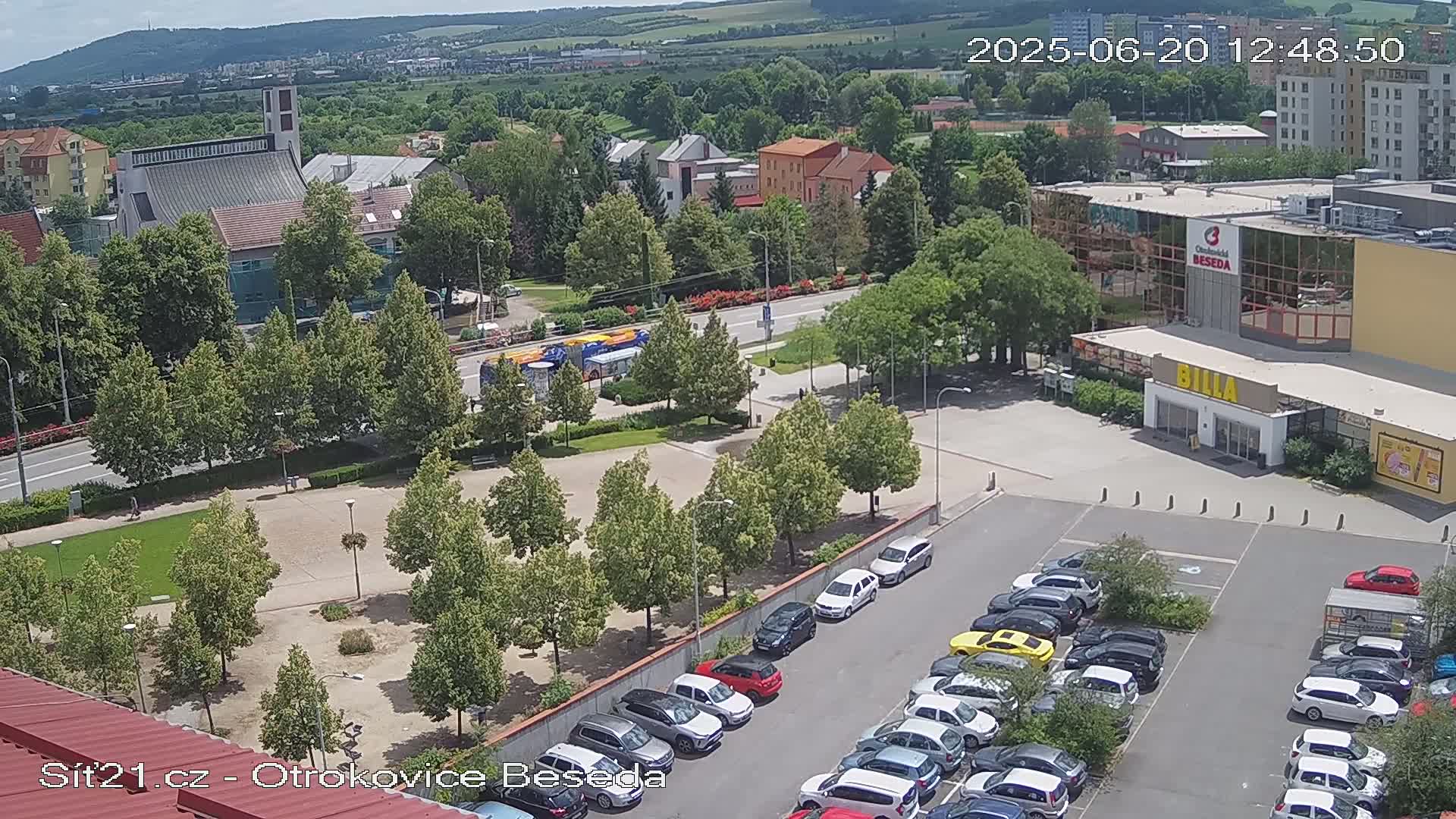 An aerial view shows a town square with a church, a large building, and a parking lot filled with cars, on a sunny day.