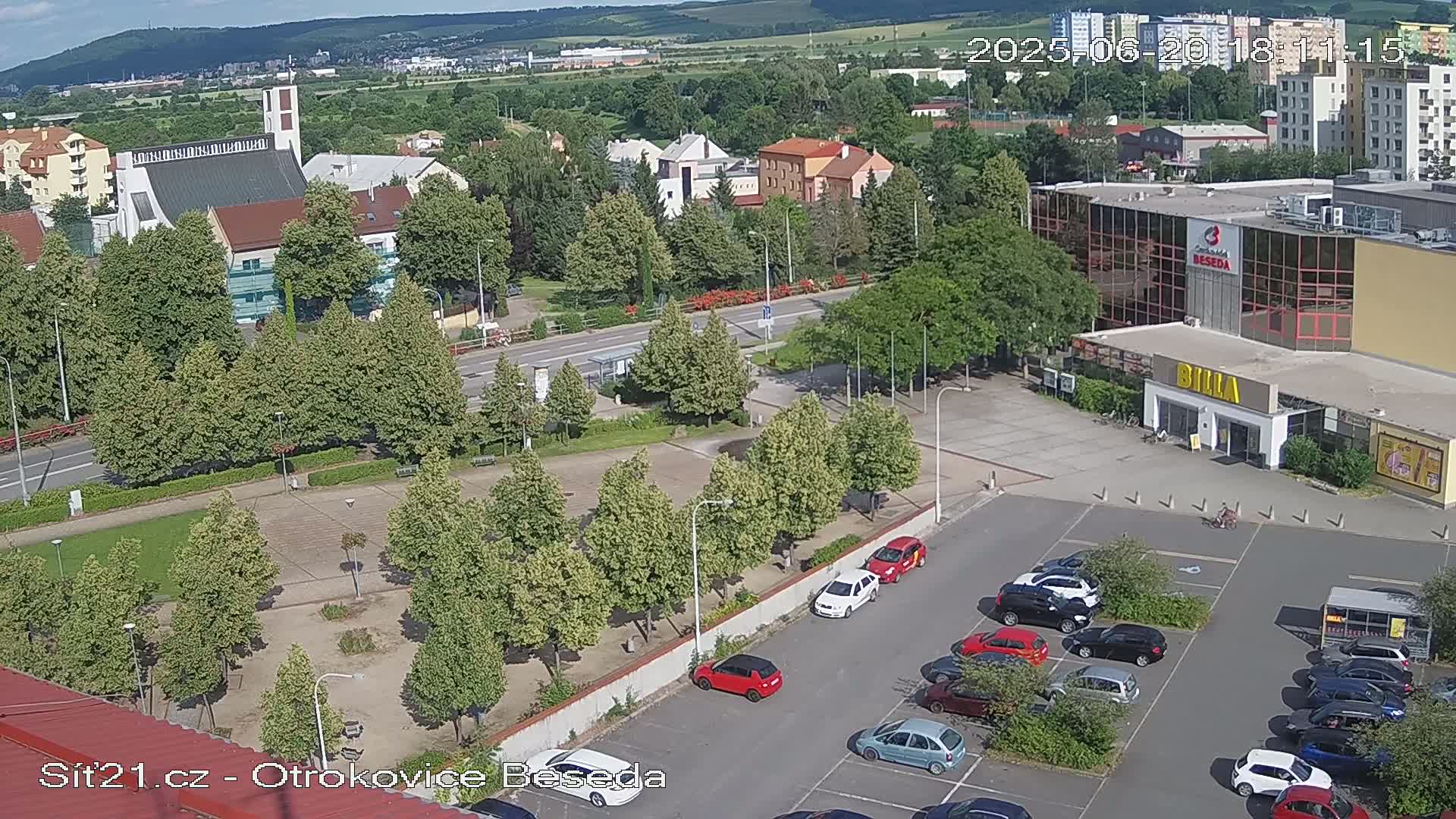 An aerial view of a town square with a church, buildings, trees, and a parking lot filled with cars on a sunny day.