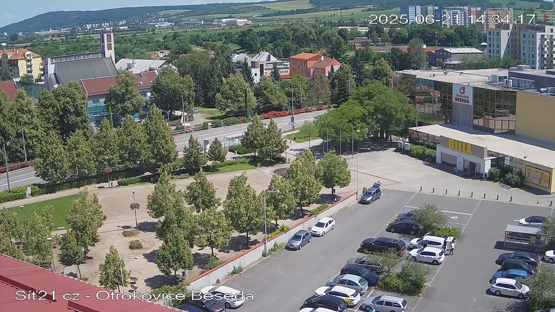 An aerial view shows a town square with green trees, a church, a large building with a parking lot full of cars, and a sunny day.