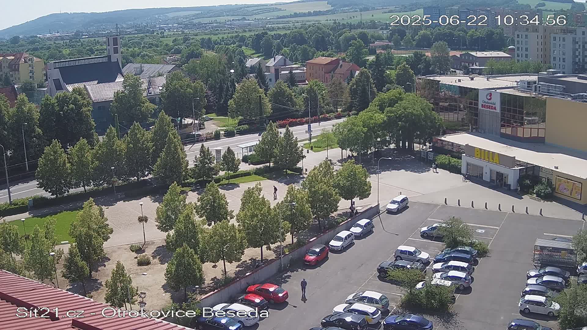 An aerial view shows a town square with a church, a supermarket, and a parking lot filled with cars on a sunny day.