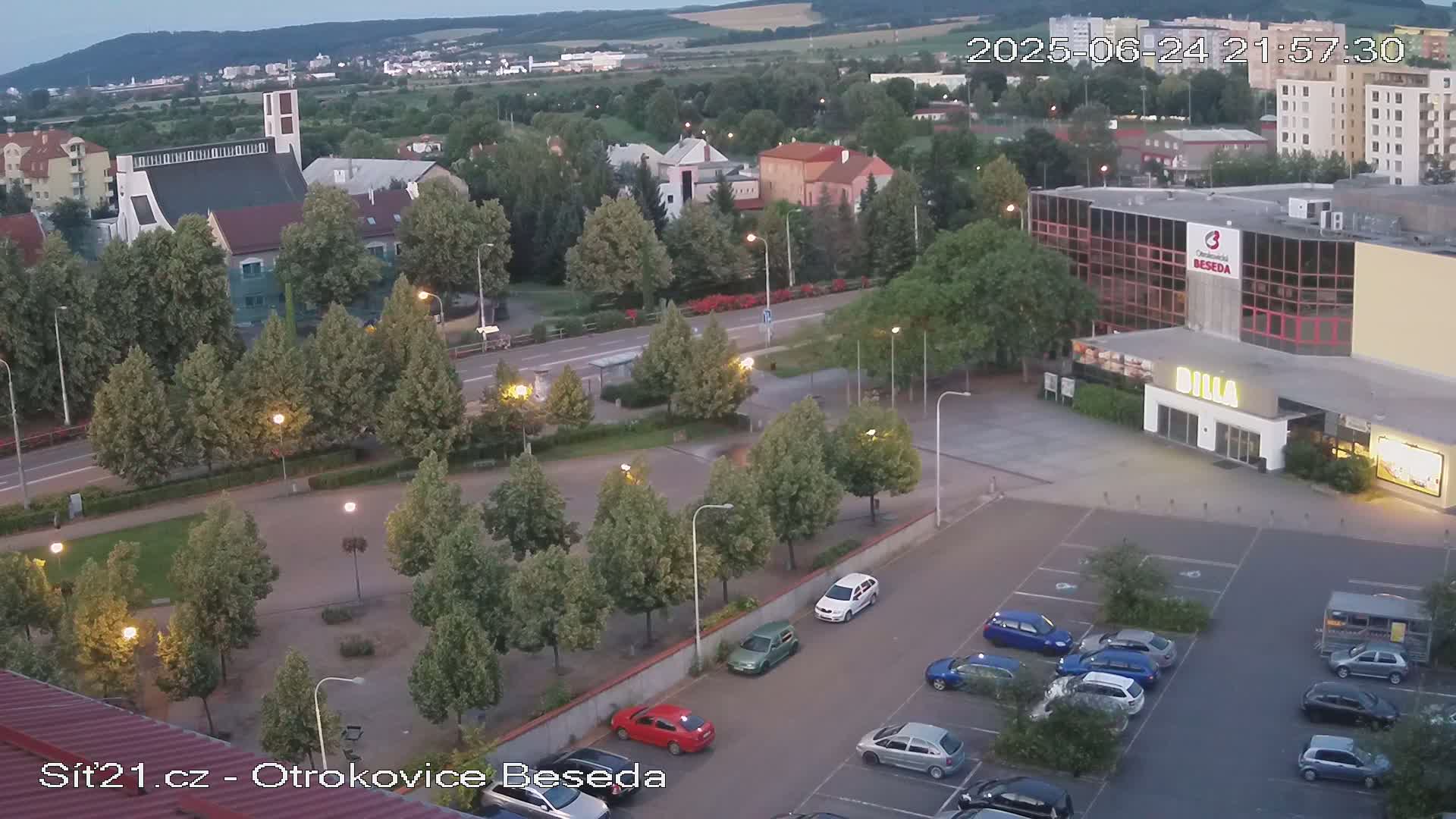 An aerial view shows a town square with a church, shops, and a parking lot at dusk under a clear sky.