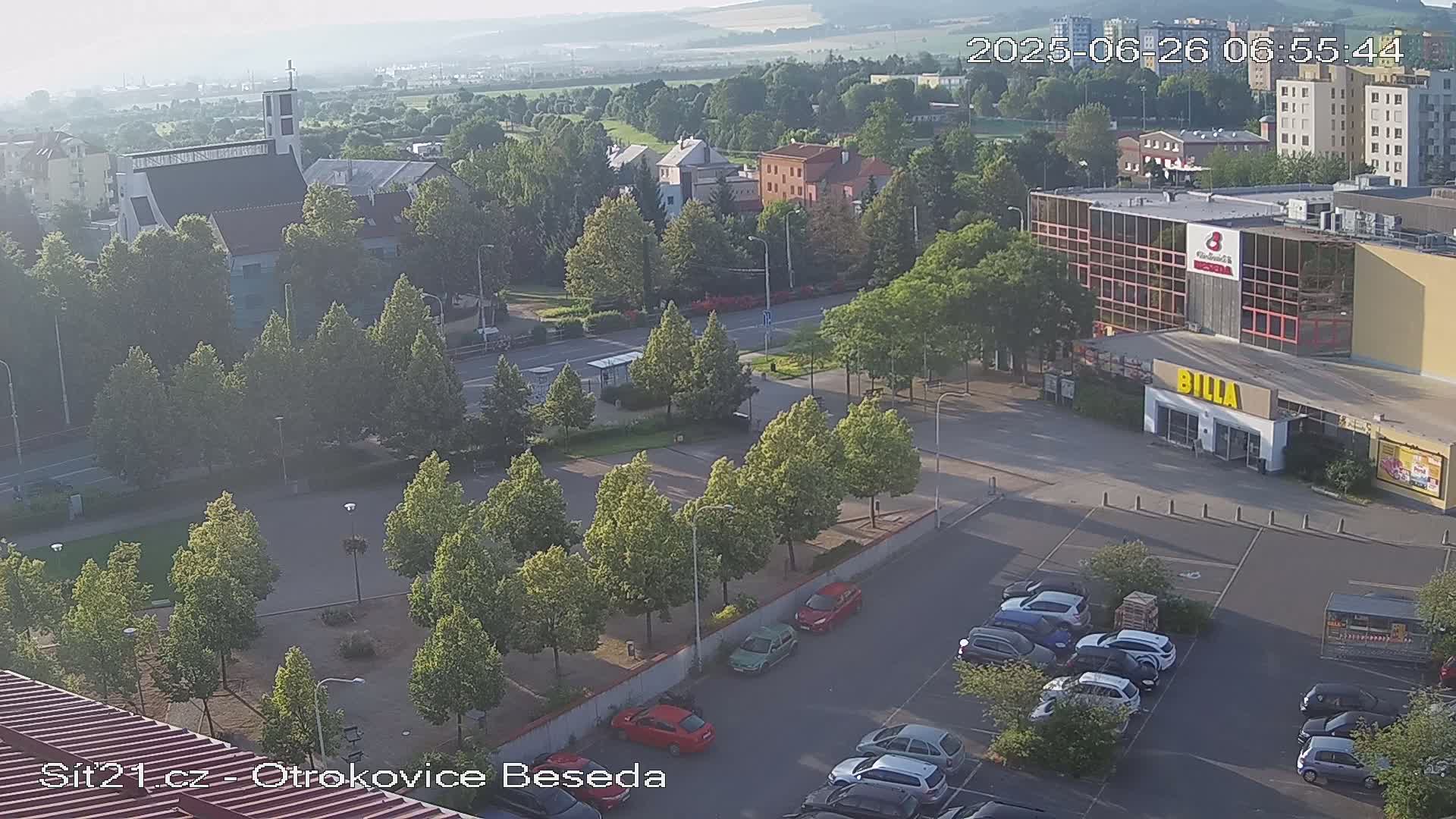 An aerial view shows a town square with a church, shops, and a parking lot at dusk under a clear sky.