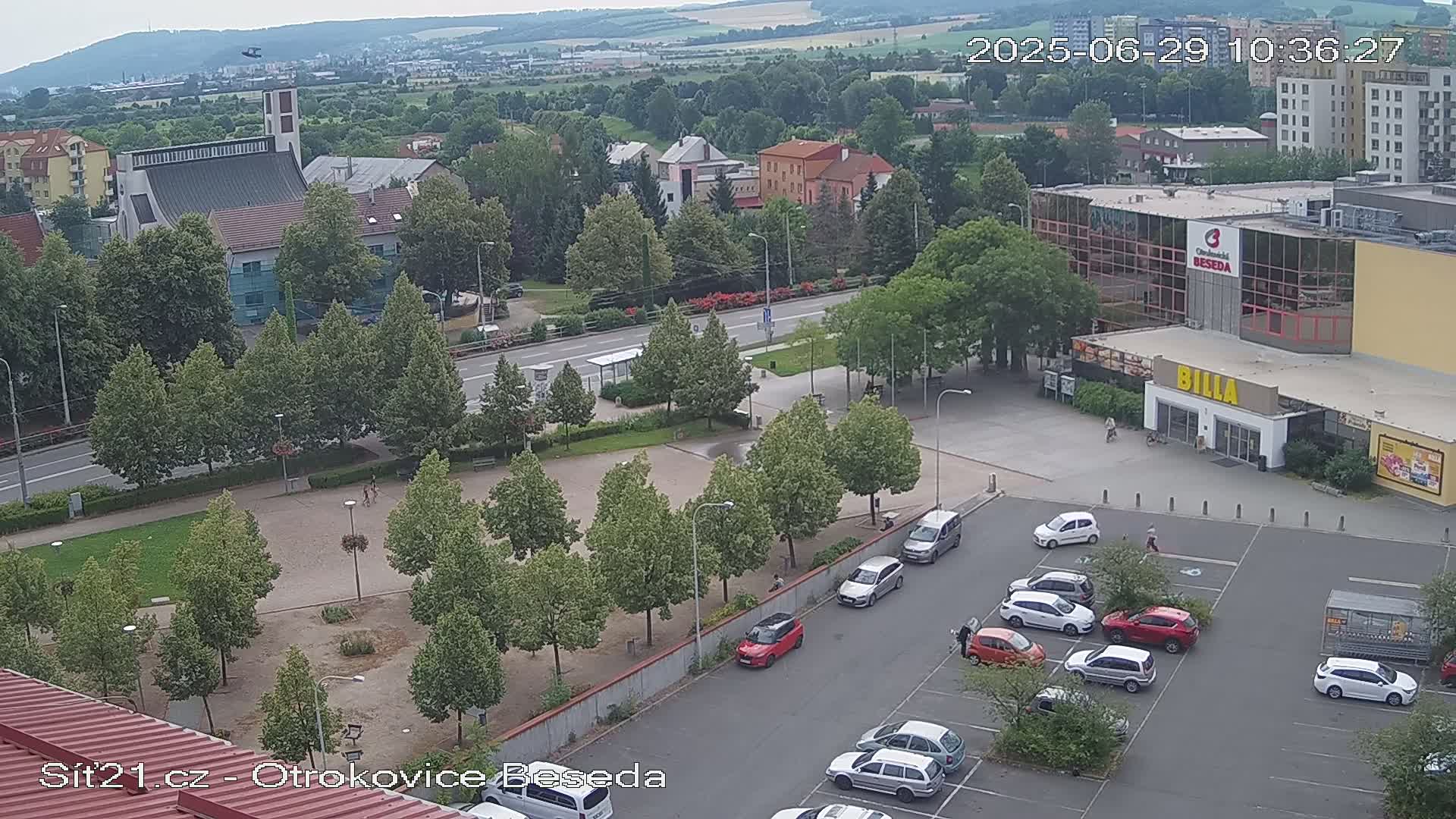 An aerial view of a town square with a church, buildings, a parking lot with cars, and green spaces under a mostly clear sky.