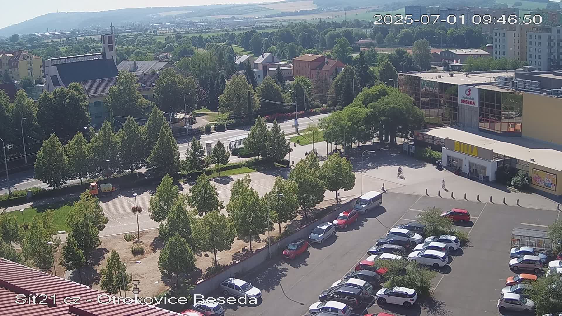 A high-angle view of a sunny parking lot next to a church and a supermarket, surrounded by trees and buildings under a clear sky.