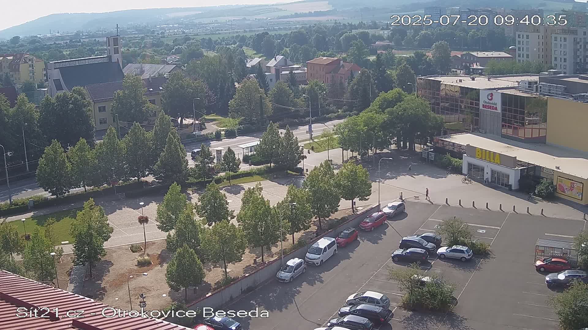 An aerial view of a town square with a parking lot, buildings, and numerous green trees on a sunny day.