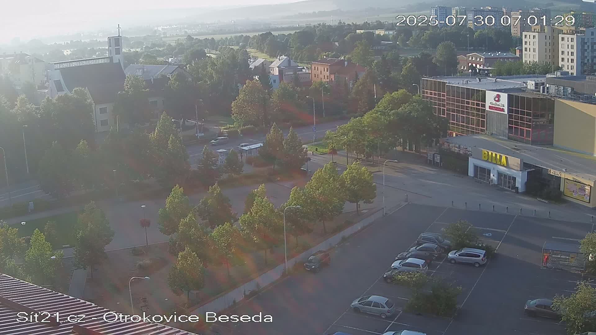 An aerial view of a town square with a parking lot, buildings, and numerous green trees on a sunny day.