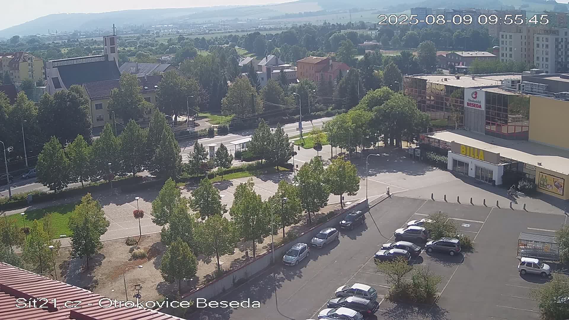 An elevated view shows a town square with a parking lot, several buildings including a church, and green spaces under a hazy, sunny sky.