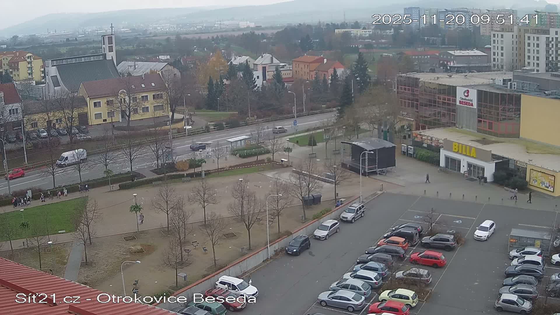 An overcast and damp day reveals an aerial view of a town featuring a prominent church, a multi-lane road with moving vehicles, numerous bare trees, pedestrians walking on paths, various buildings including a supermarket, and a large parking lot filled with cars, all set against hazy distant hills.