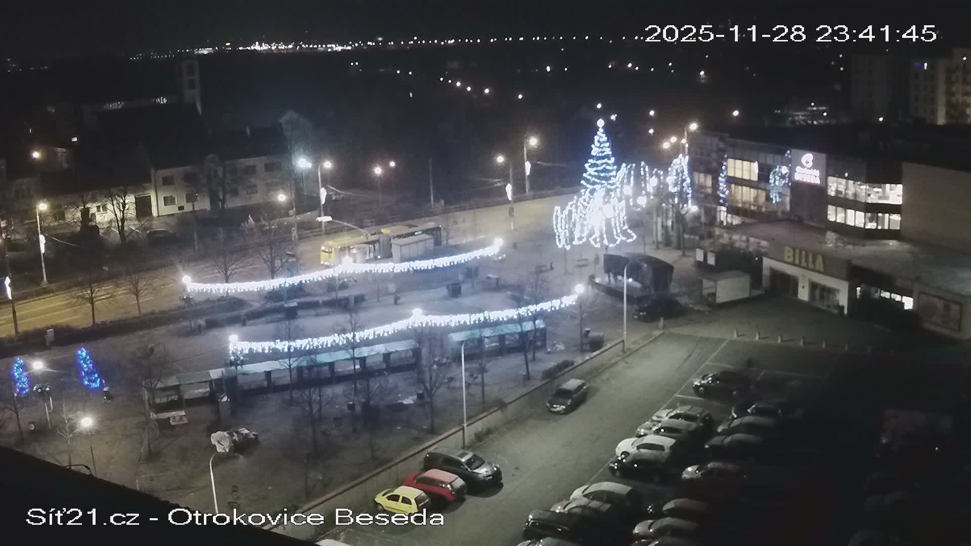 A clear night view captures an urban square illuminated by festive white string lights and a large Christmas tree, surrounded by buildings, a street, and a parking lot filled with cars.