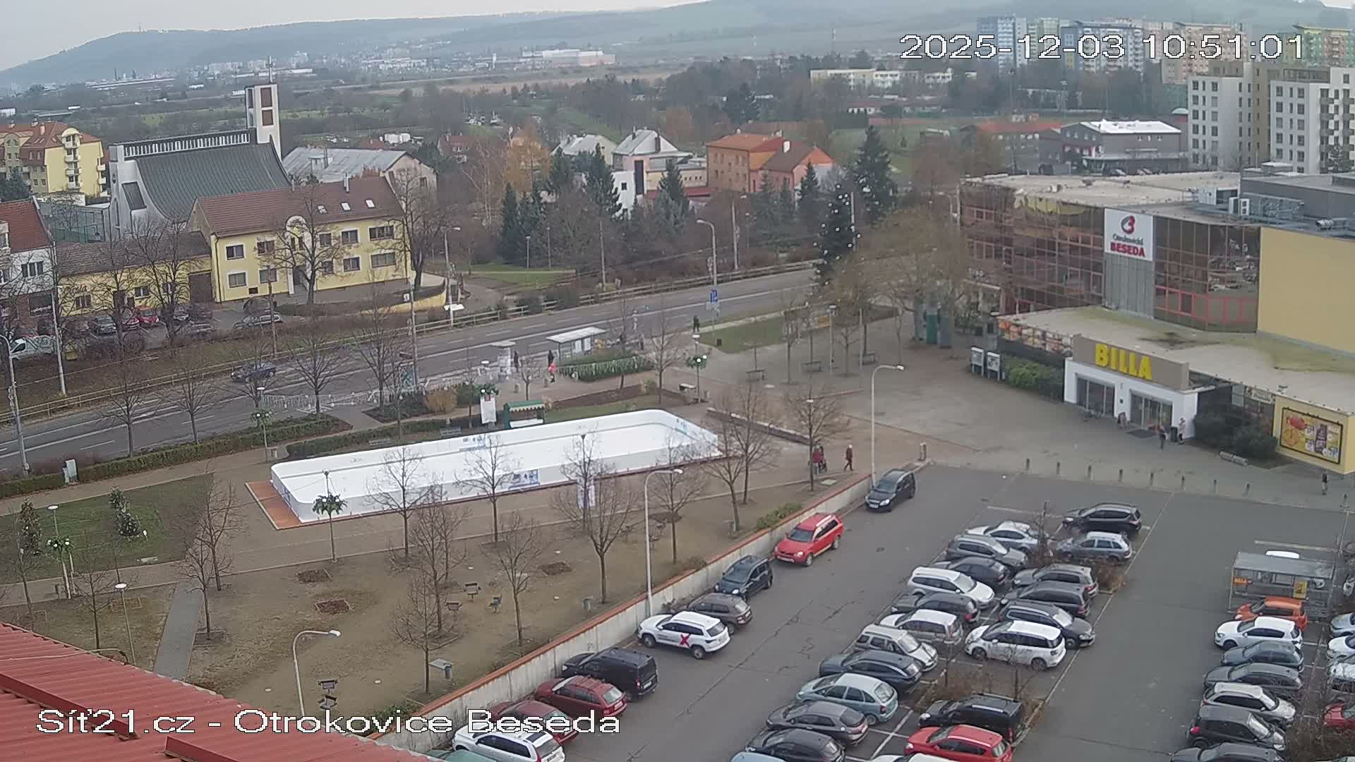 An overcast, high-angle view of a bustling urban area features an outdoor ice rink, numerous buildings including a church and a supermarket, a busy road, and a large parking lot filled with cars.