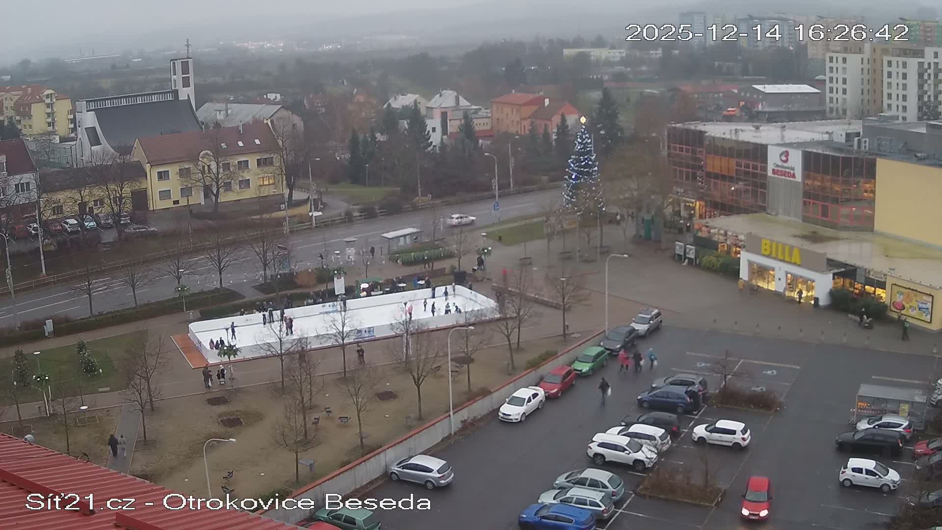 An overcast, high-angle view of a bustling urban area features an outdoor ice rink, numerous buildings including a church and a supermarket, a busy road, and a large parking lot filled with cars.