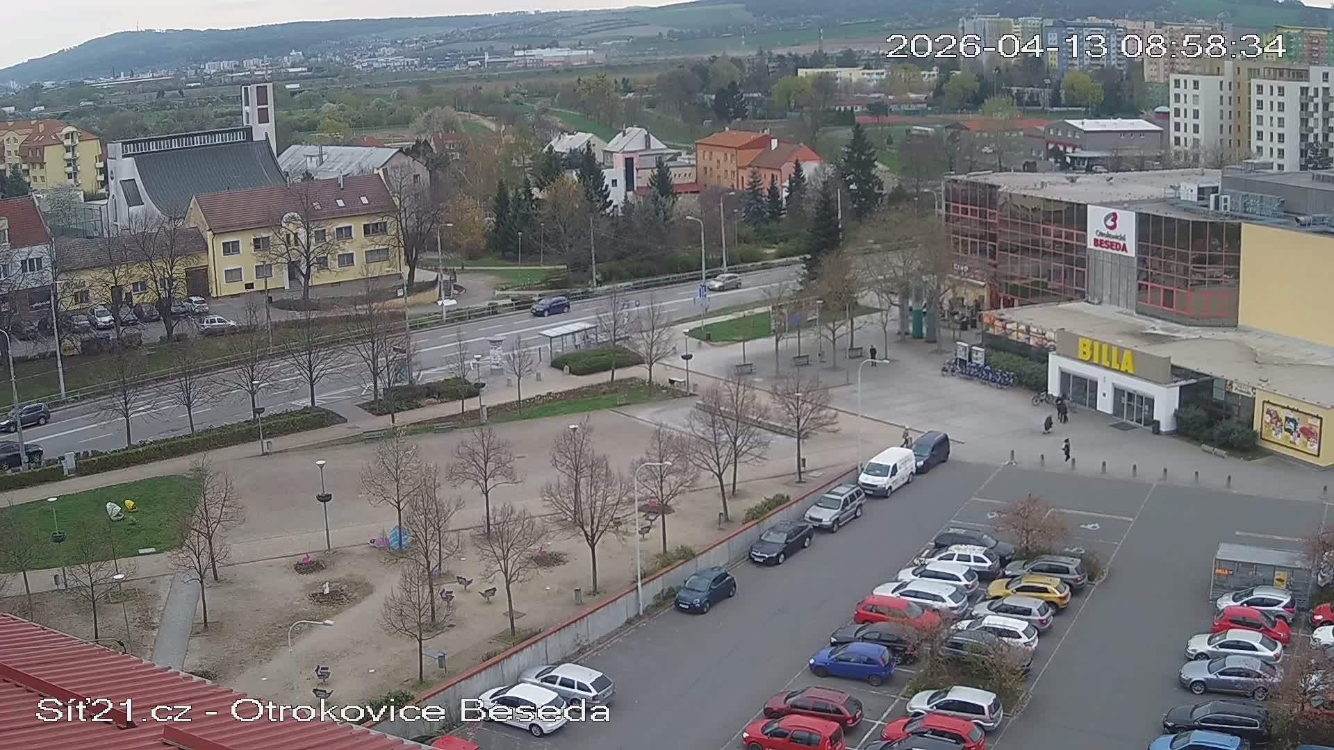 An overcast, high-angle view of a bustling urban area features an outdoor ice rink, numerous buildings including a church and a supermarket, a busy road, and a large parking lot filled with cars.