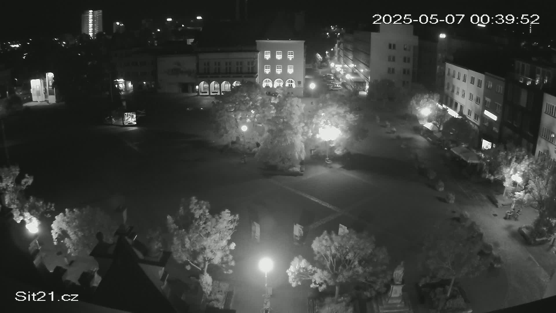 A nighttime aerial view of a town square shows several buildings, trees, and streetlights illuminating the area.