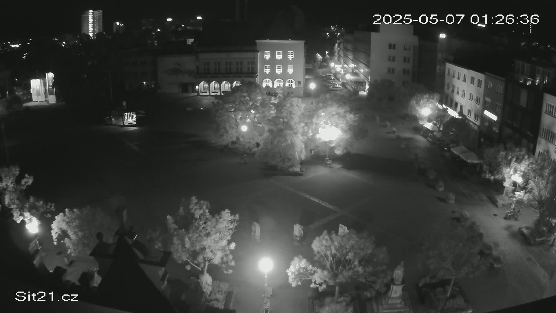 A nighttime, high-angle view of a town square with buildings surrounding it, illuminated by streetlights and featuring several trees and sparsely populated with people.