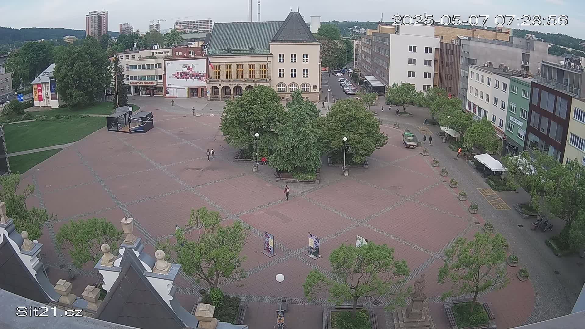 An overcast day reveals a brick town square with several buildings surrounding it, featuring trees, benches, and a few pedestrians.