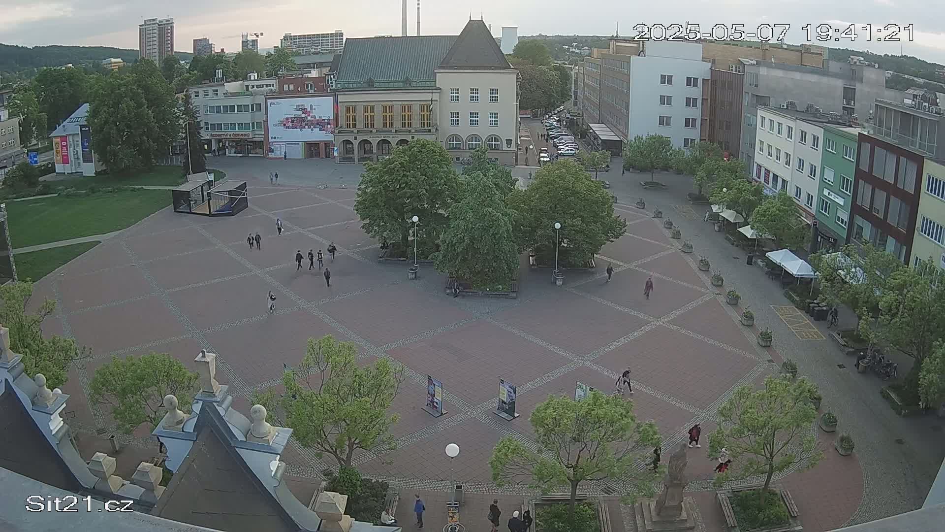 A high-angle view shows a town square with several people walking around, under an overcast sky.