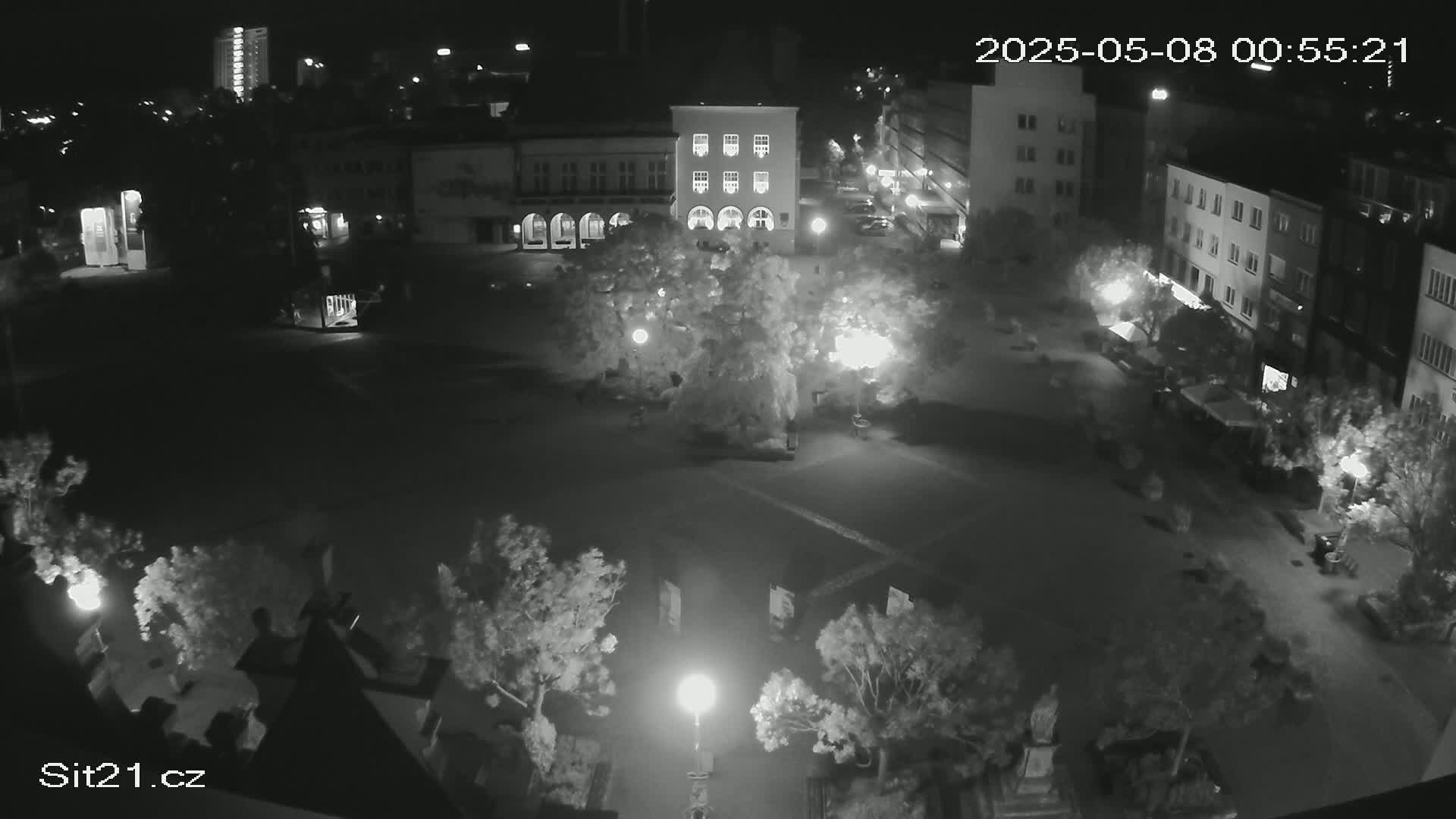 A nighttime, high-angle view of a town square illuminated by streetlights, featuring buildings, trees, and sparsely populated pathways.