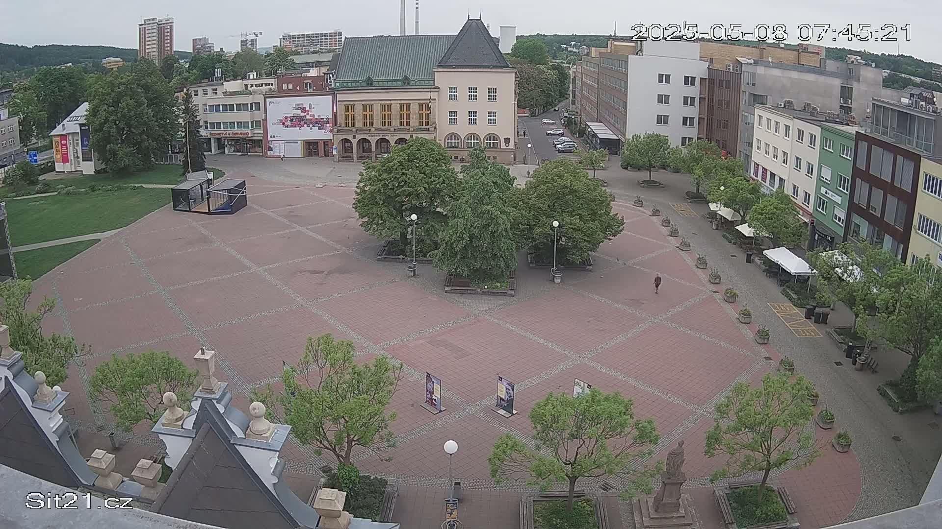 An overcast day reveals a mostly empty town square with several buildings surrounding it, small trees and benches dotting the area, and a statue in the bottom right corner.