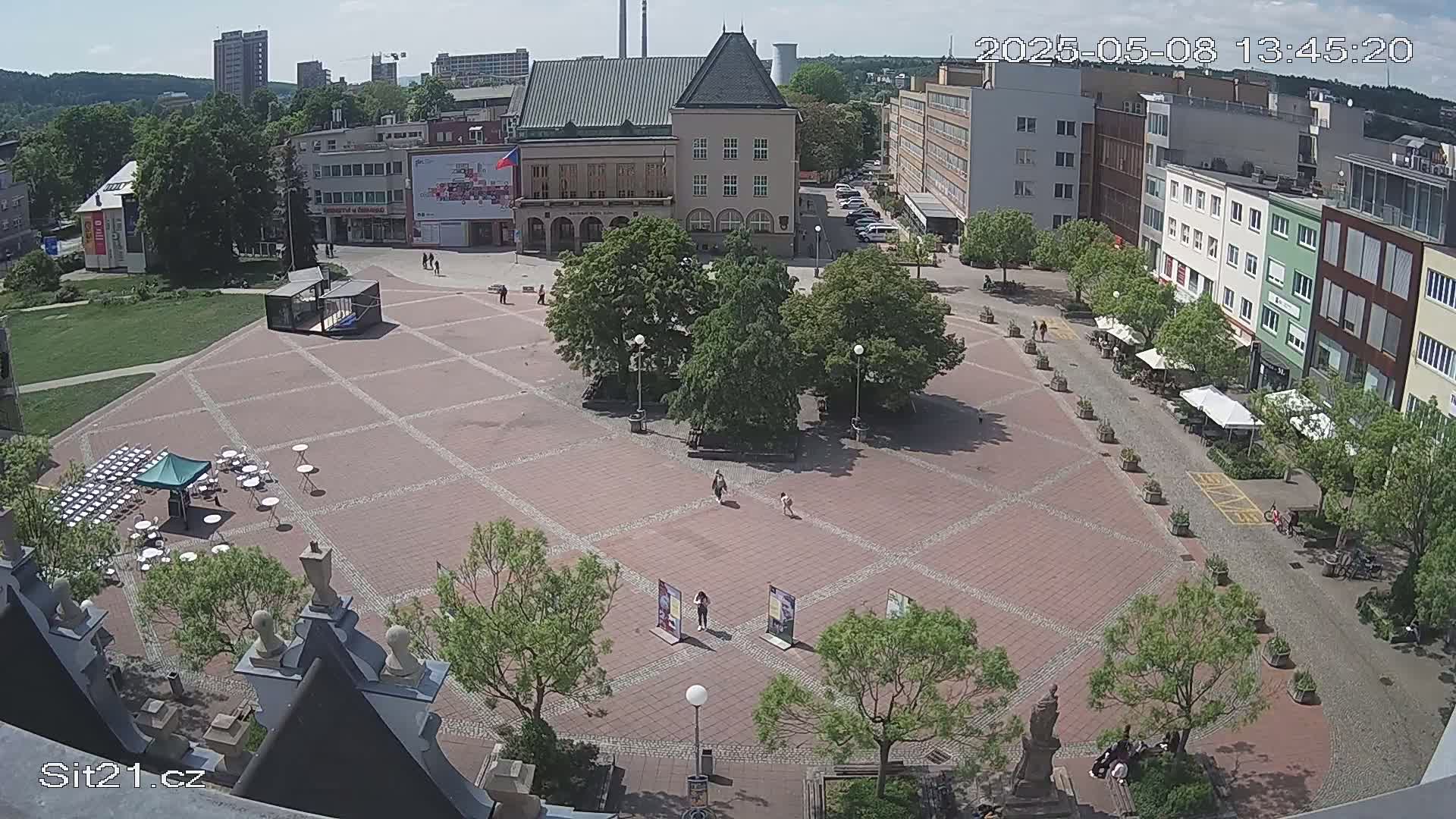 A sunny day reveals a brick town square with several buildings surrounding it, some trees, and a few people walking around.