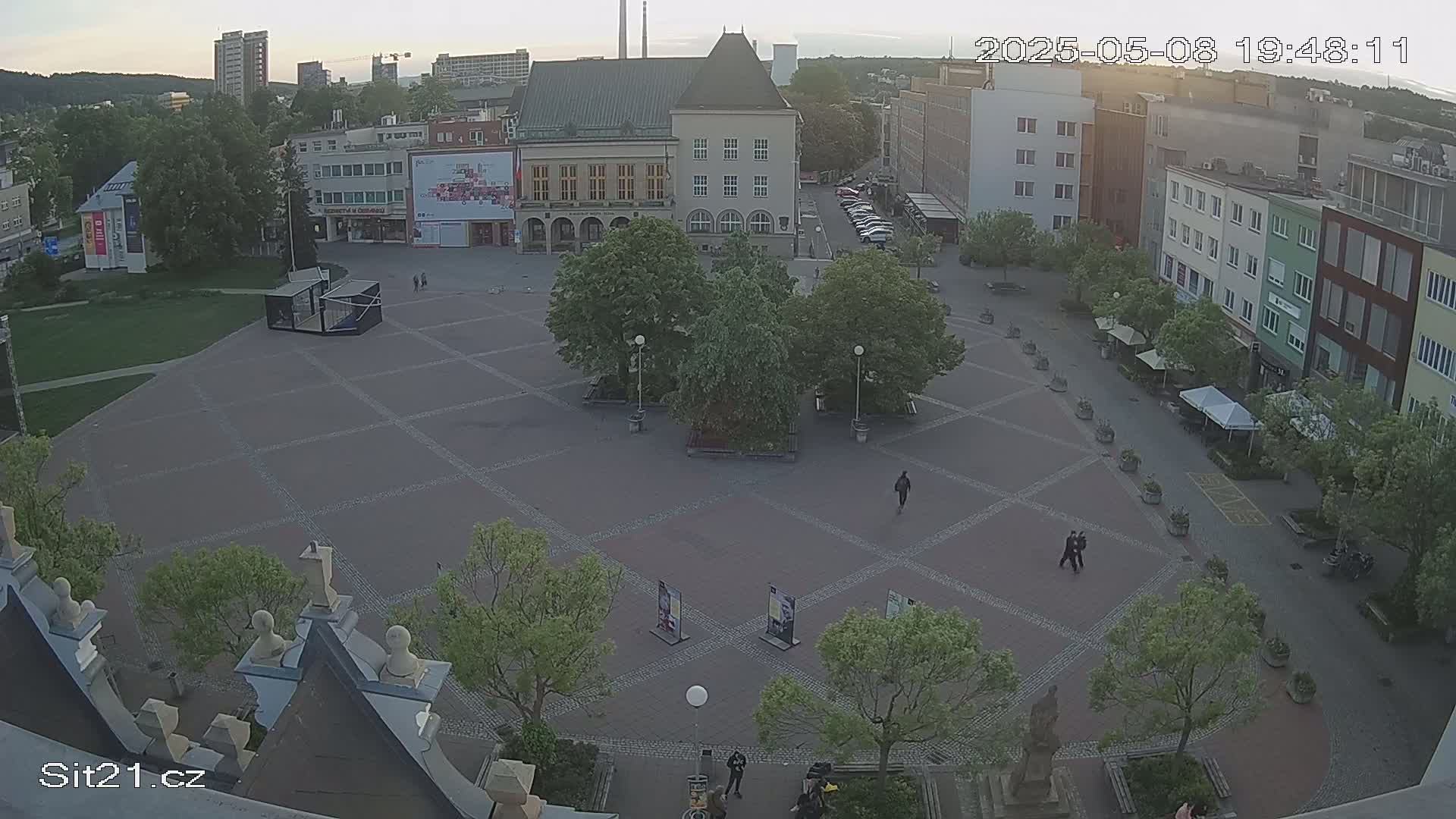 A paved town square, partially shaded by trees, is sparsely populated with a few pedestrians under a dusky sky.