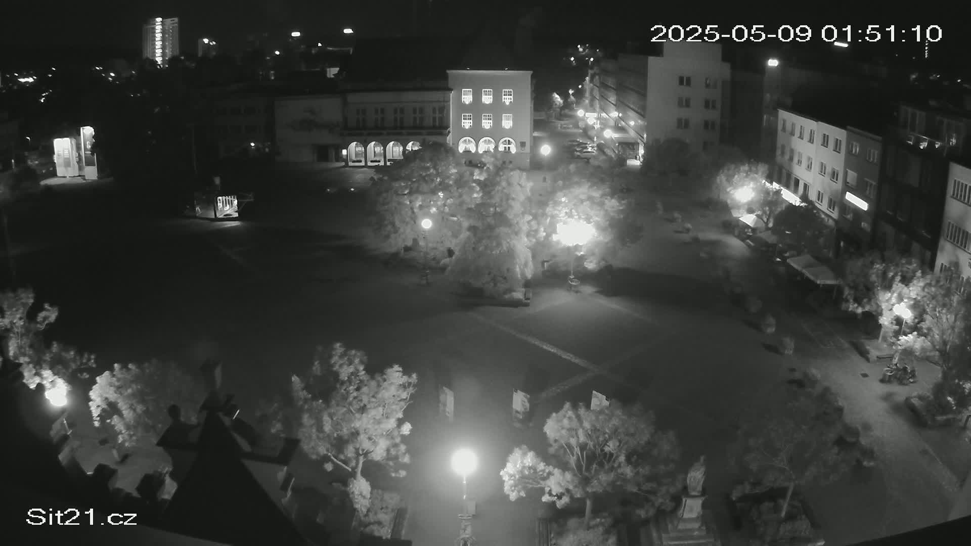 A nighttime, high-angle view of a town square with several buildings, trees, and streetlights illuminated, under a clear sky.
