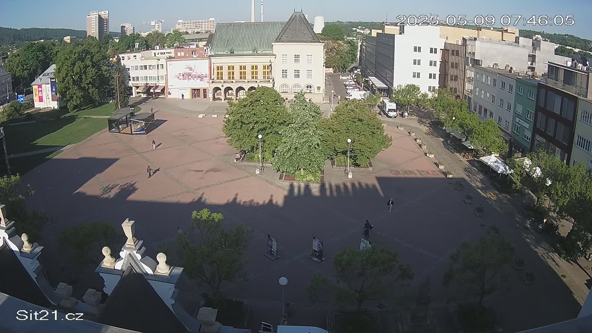 A mostly empty town square is seen on a sunny day, with several buildings surrounding it, including a large building with a pitched roof and some small trees and seating areas within the square.
