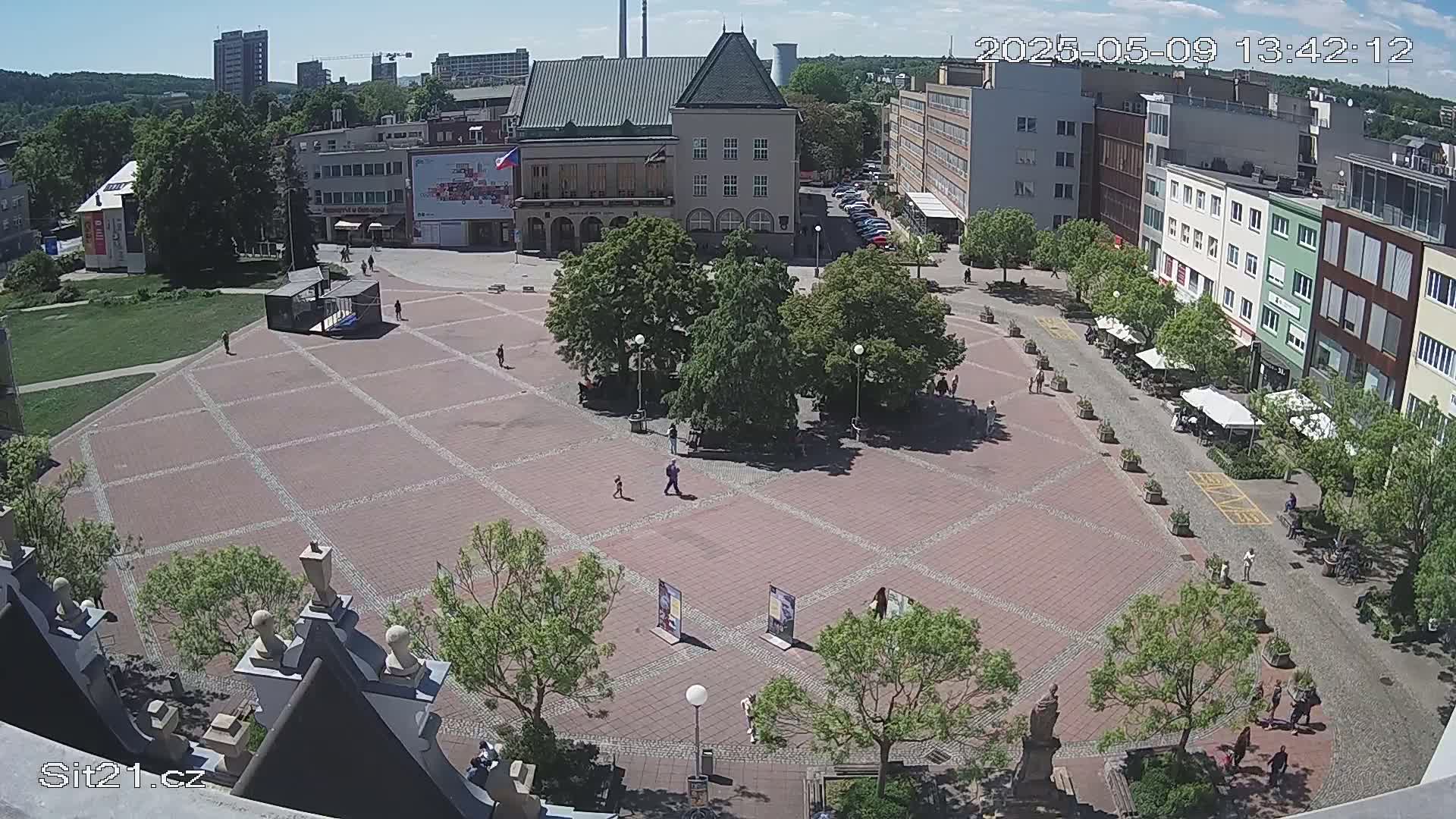 A brick town square with several buildings surrounding it, dotted with trees and a few people, on a sunny day.
