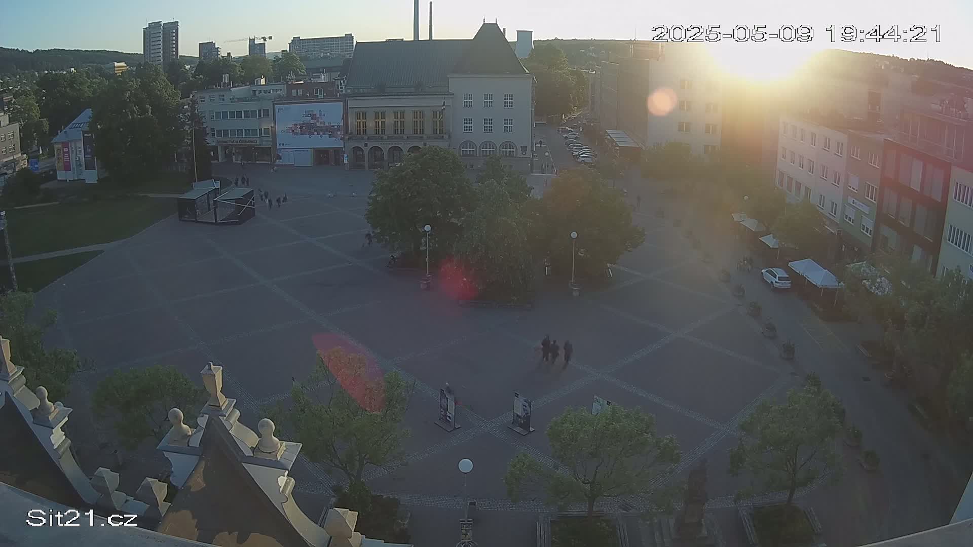An aerial view of a town square at sunset, showing several buildings, a central tree, and a few pedestrians.