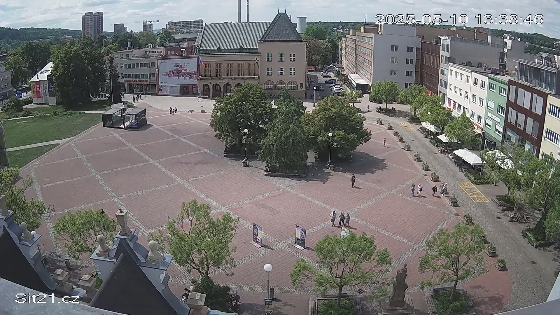 A large brick town square with several buildings surrounding it, a few people walking around, and trees providing shade under a partly sunny sky.
