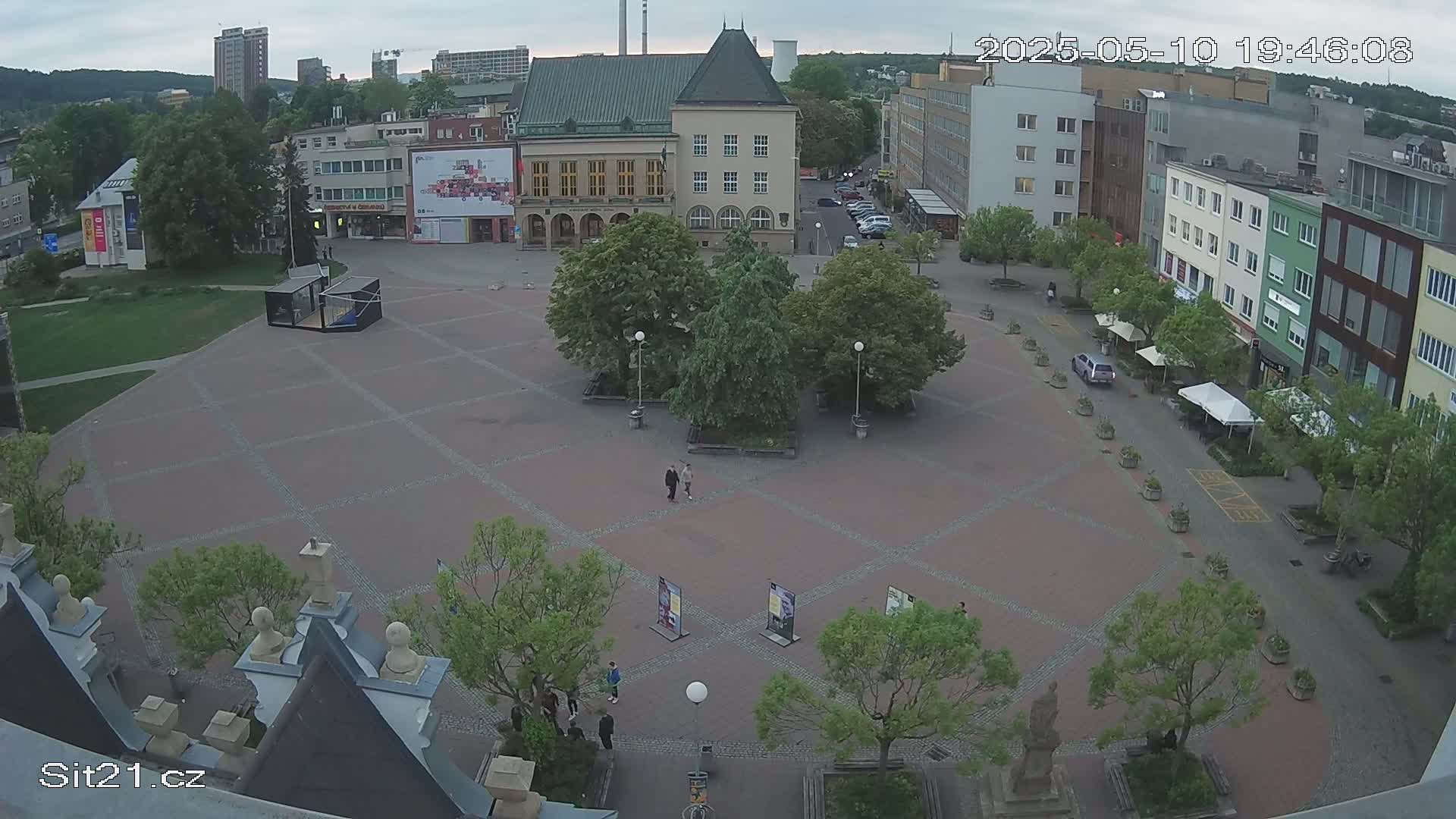 An overcast day reveals a town square with several buildings, green spaces, and a few people walking.