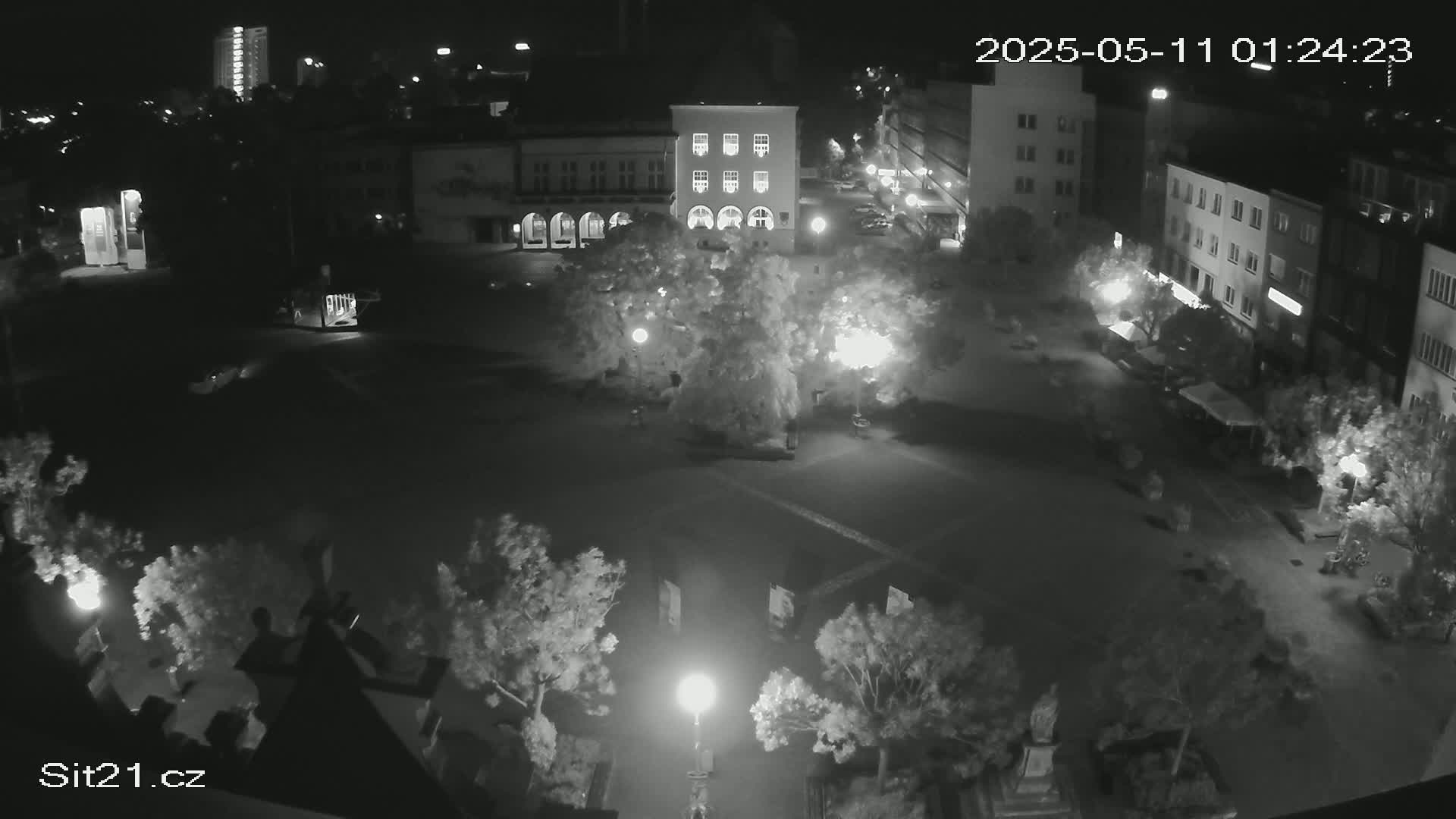 A nighttime, black-and-white overhead view of a town square with several buildings, trees, and streetlights.