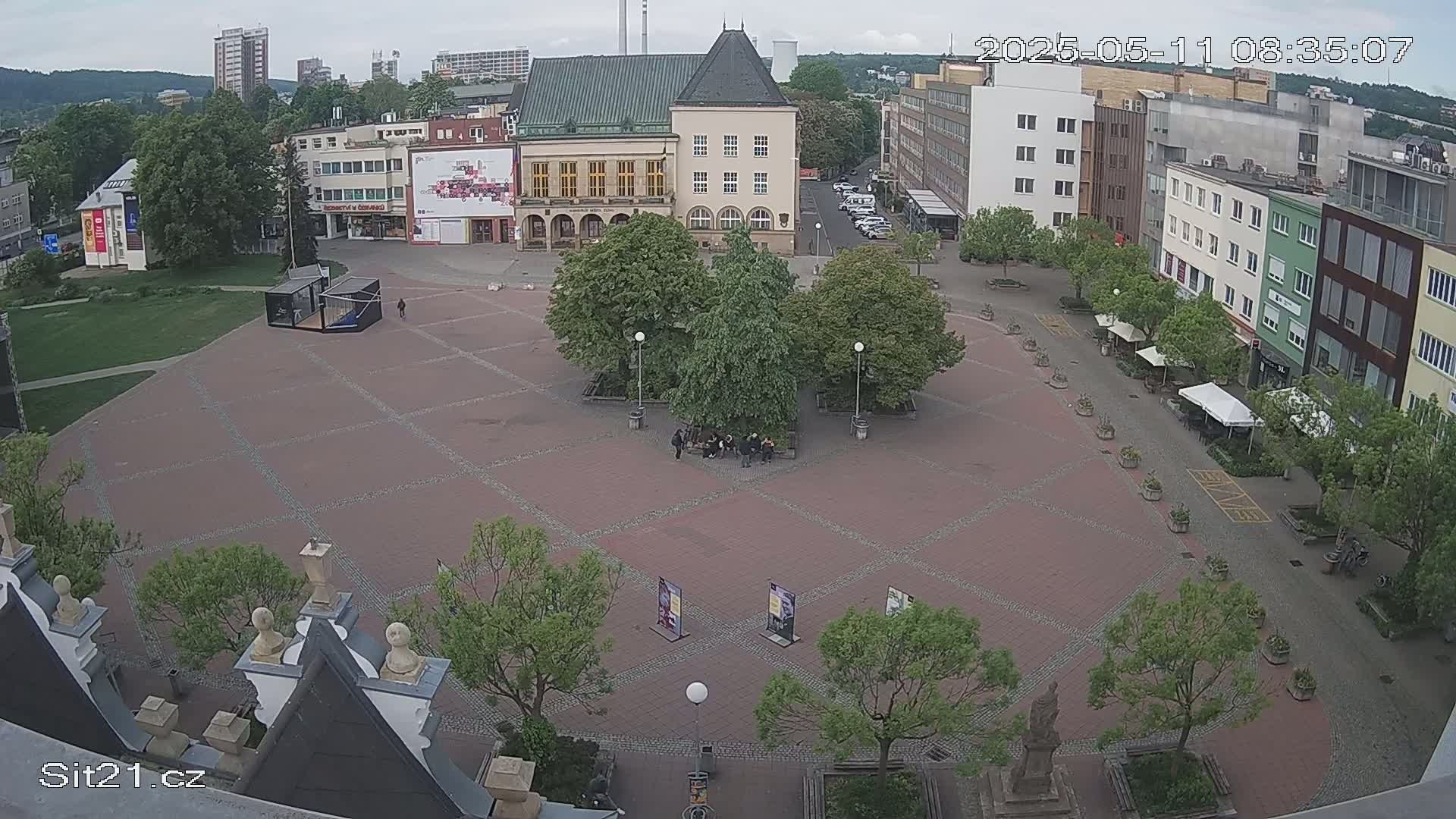 An overcast day in a town square with brick paving, several buildings, and green trees.