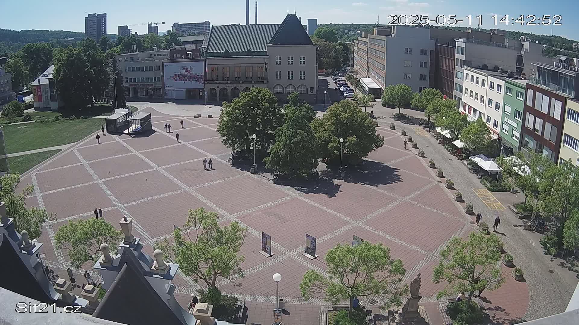 A large, brick plaza with scattered pedestrians and several trees is surrounded by buildings under a sunny sky.