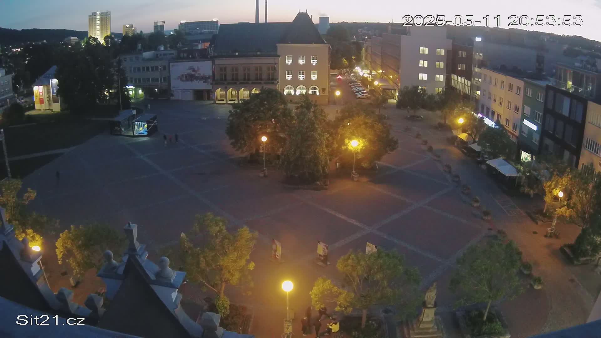 A high-angle, nighttime view of a town square with several buildings surrounding it, lit by streetlights under a twilight sky.