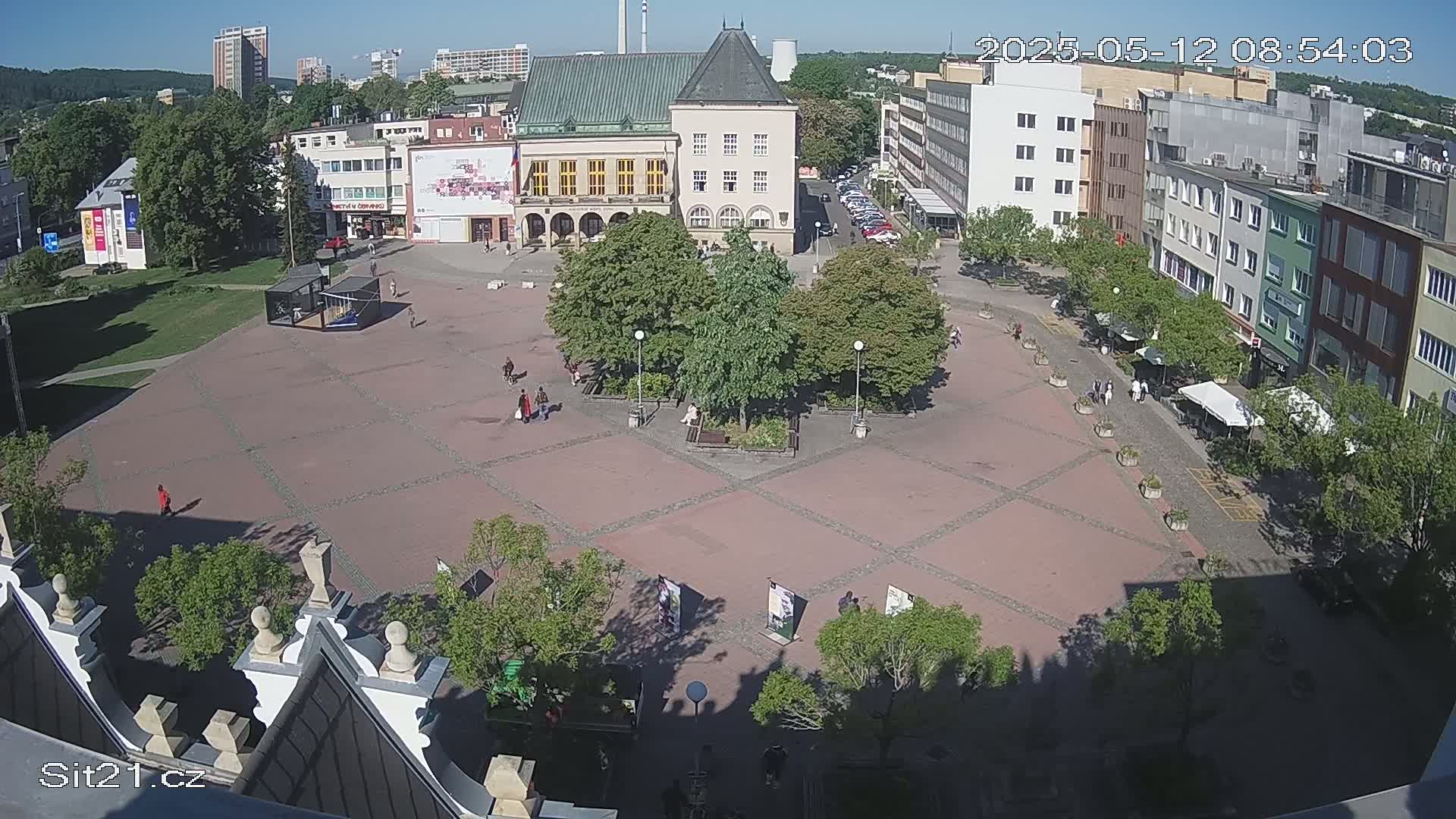 A sunny town square with several buildings surrounding it, featuring a paved area with scattered trees and people walking around.