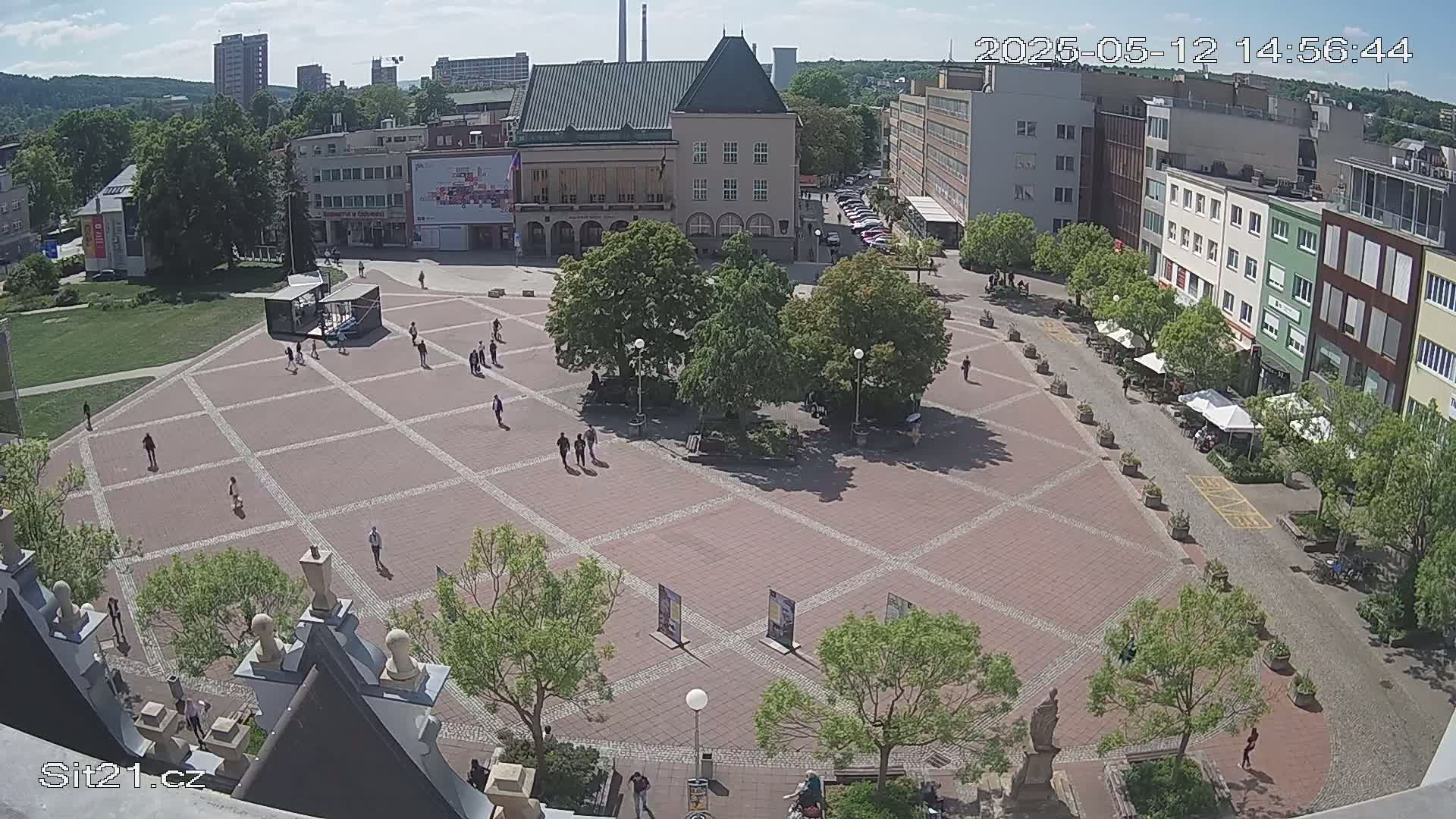 A sunny day reveals a large, brick town square with people walking among several trees and buildings surrounding it.