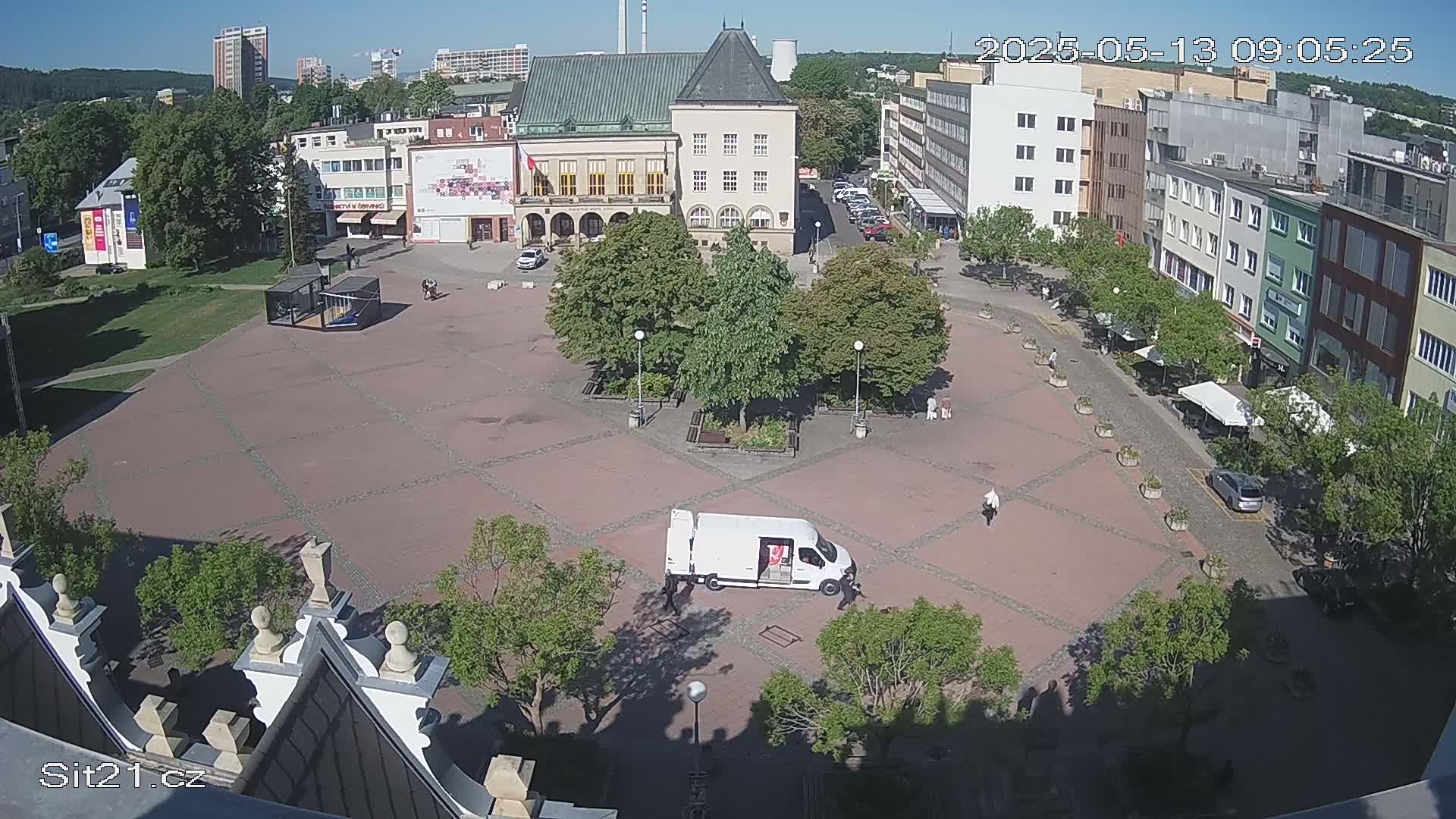 A paved town square with several buildings surrounding it, some trees, and a white van, all under a sunny sky.