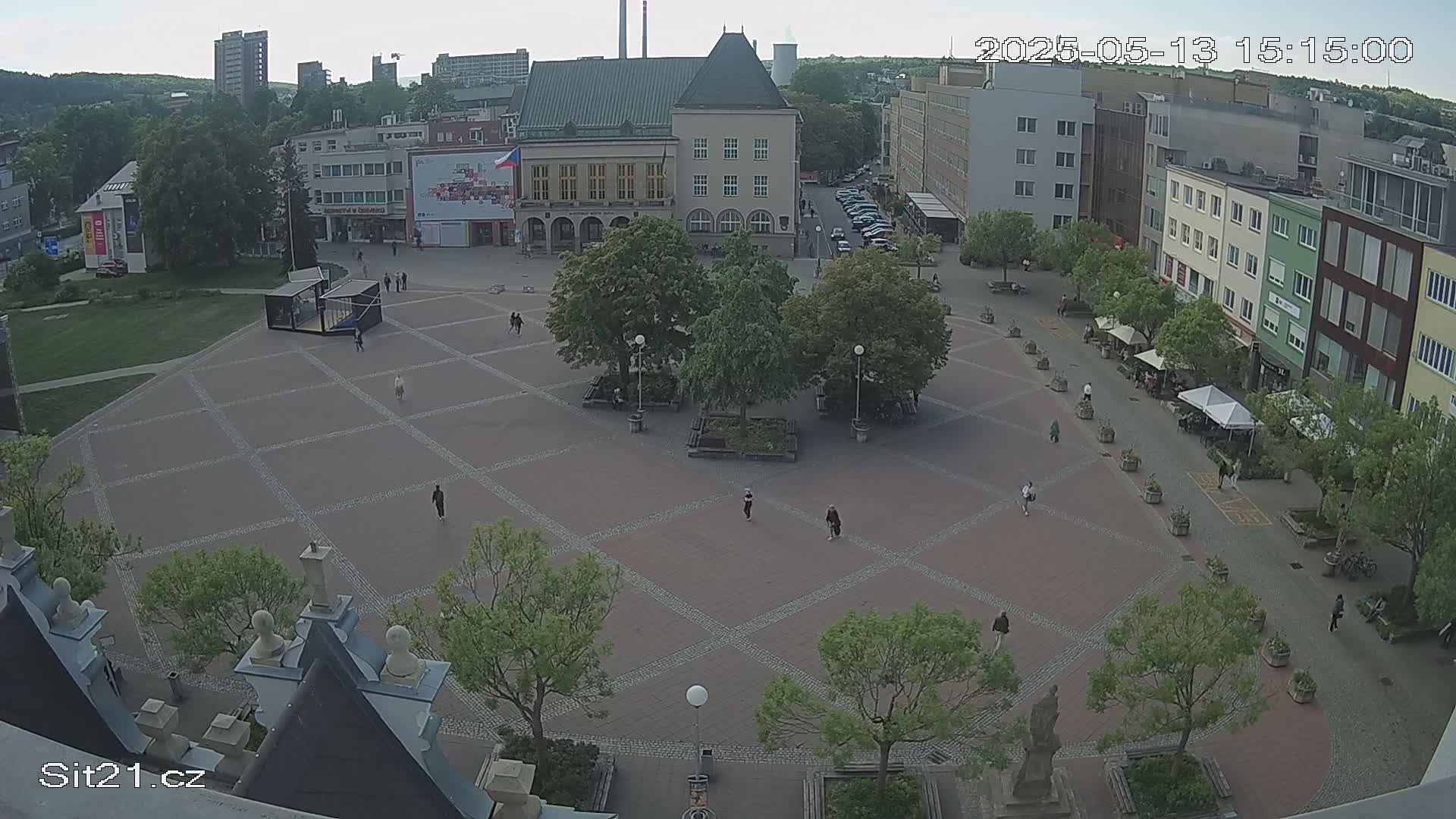 A large town square with a brick pavement, several buildings, and scattered pedestrians is visible under an overcast sky.
