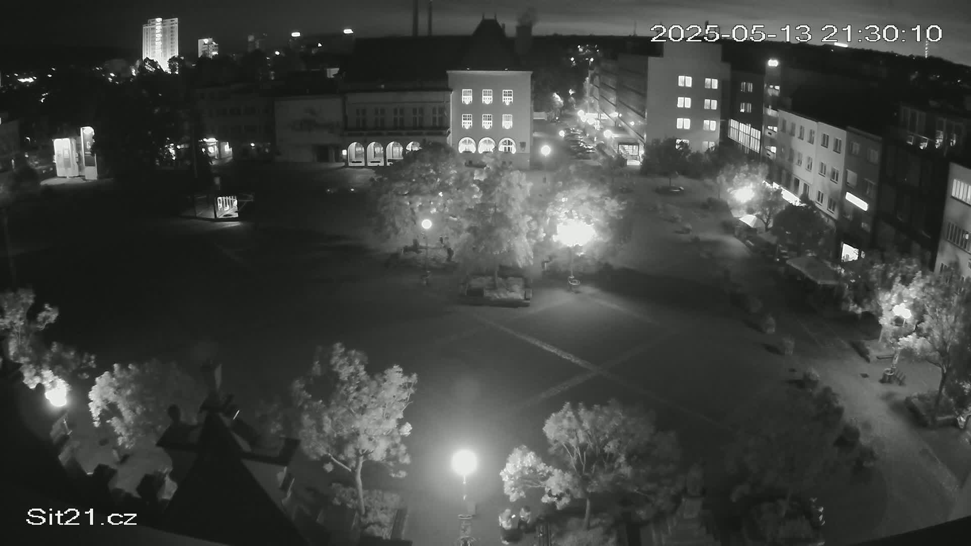 A nighttime, high-angle, black-and-white view of a town square with buildings, trees, and sparse pedestrian activity under artificial lighting.