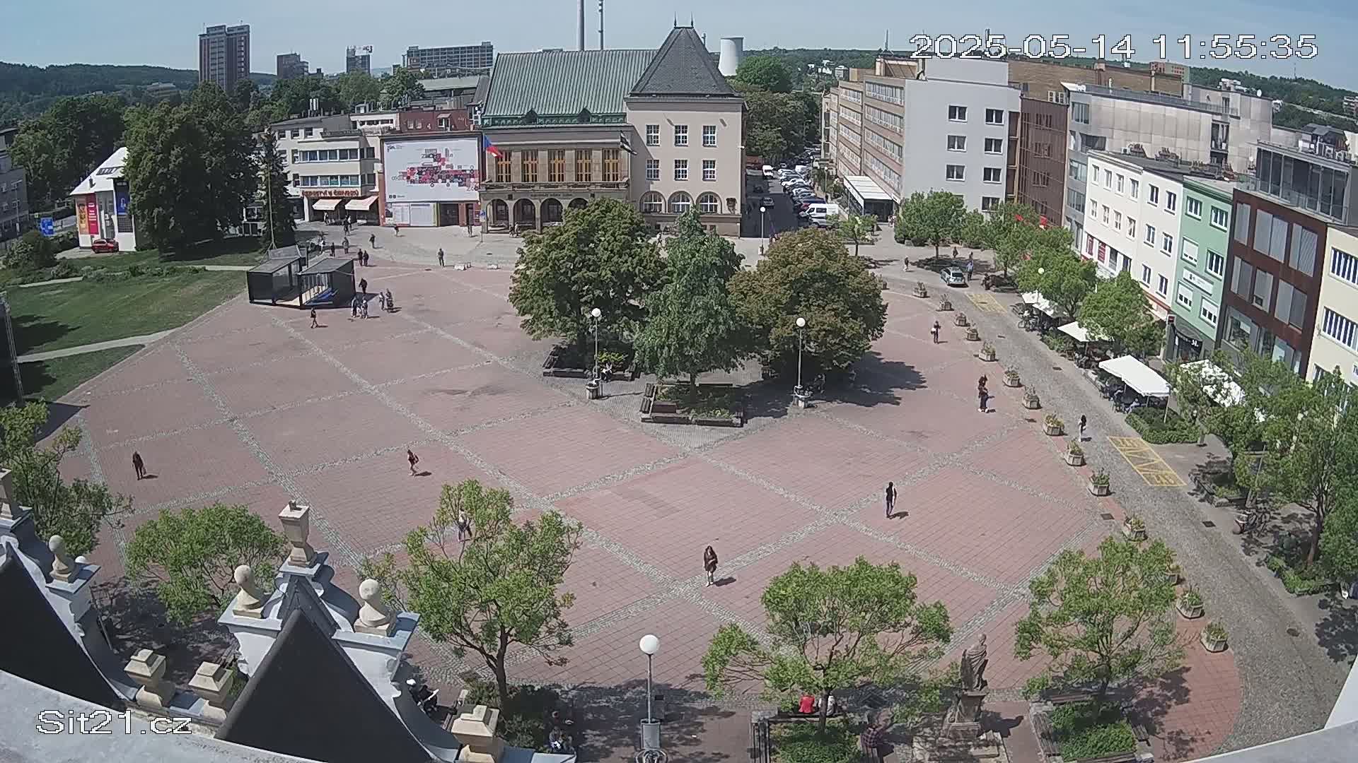 A paved town square with a few people walking around, surrounded by buildings and trees under a sunny sky.