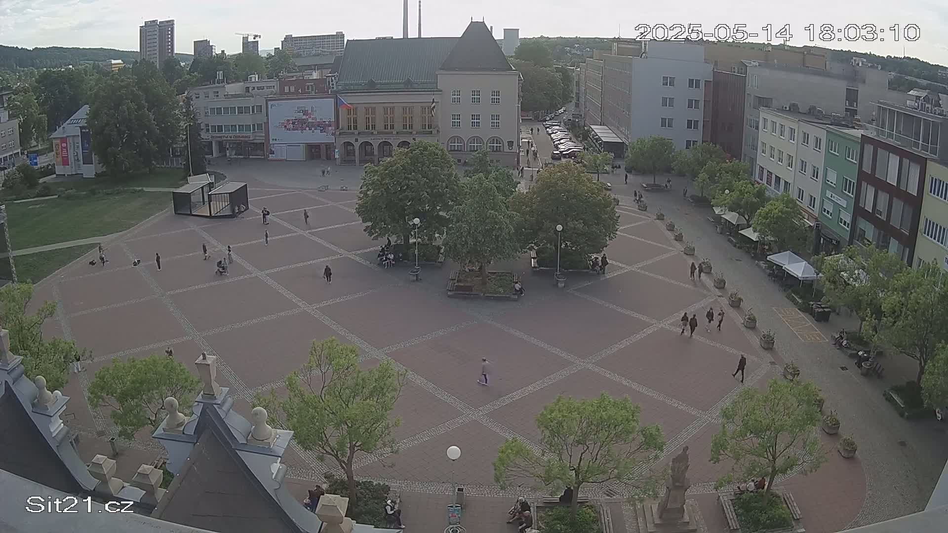 An overcast day reveals a town square with a geometric patterned pavement, surrounded by buildings and several people walking, with lush green trees providing shade.