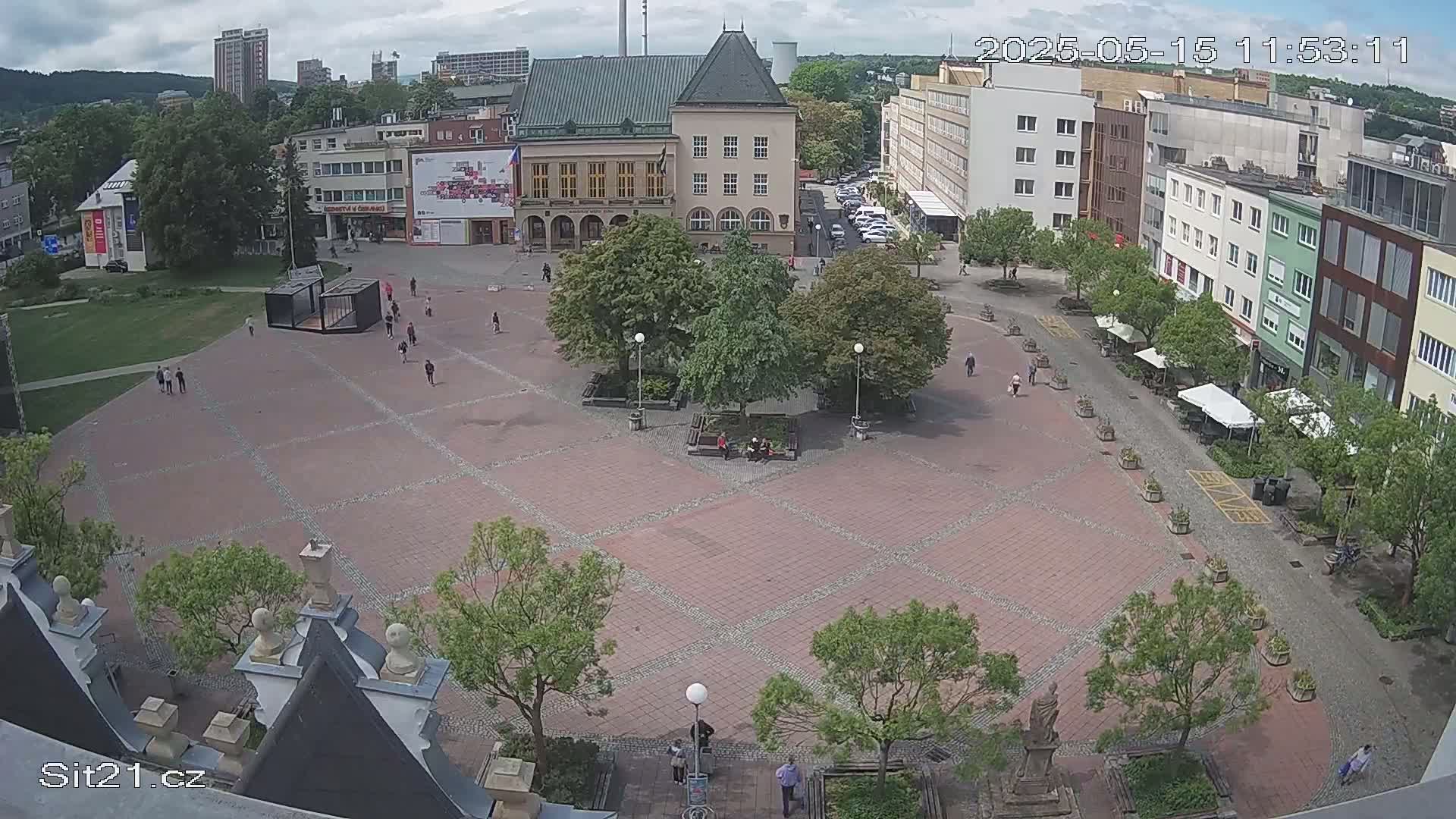 A large, brick town square with several buildings surrounding it, a few people walking around, and some trees and potted plants, under partly cloudy skies.
