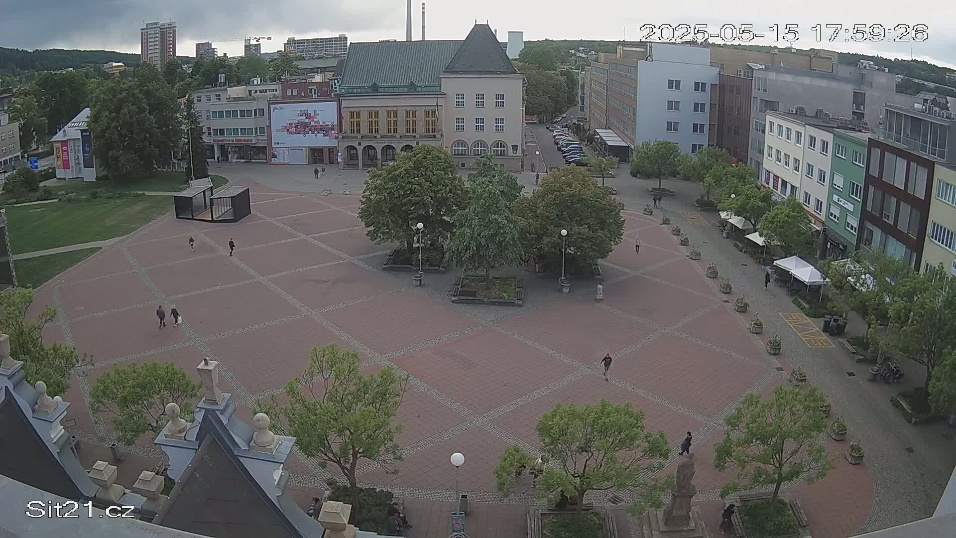 An overcast day reveals a large brick plaza with several people walking around, surrounded by buildings and lush green trees.