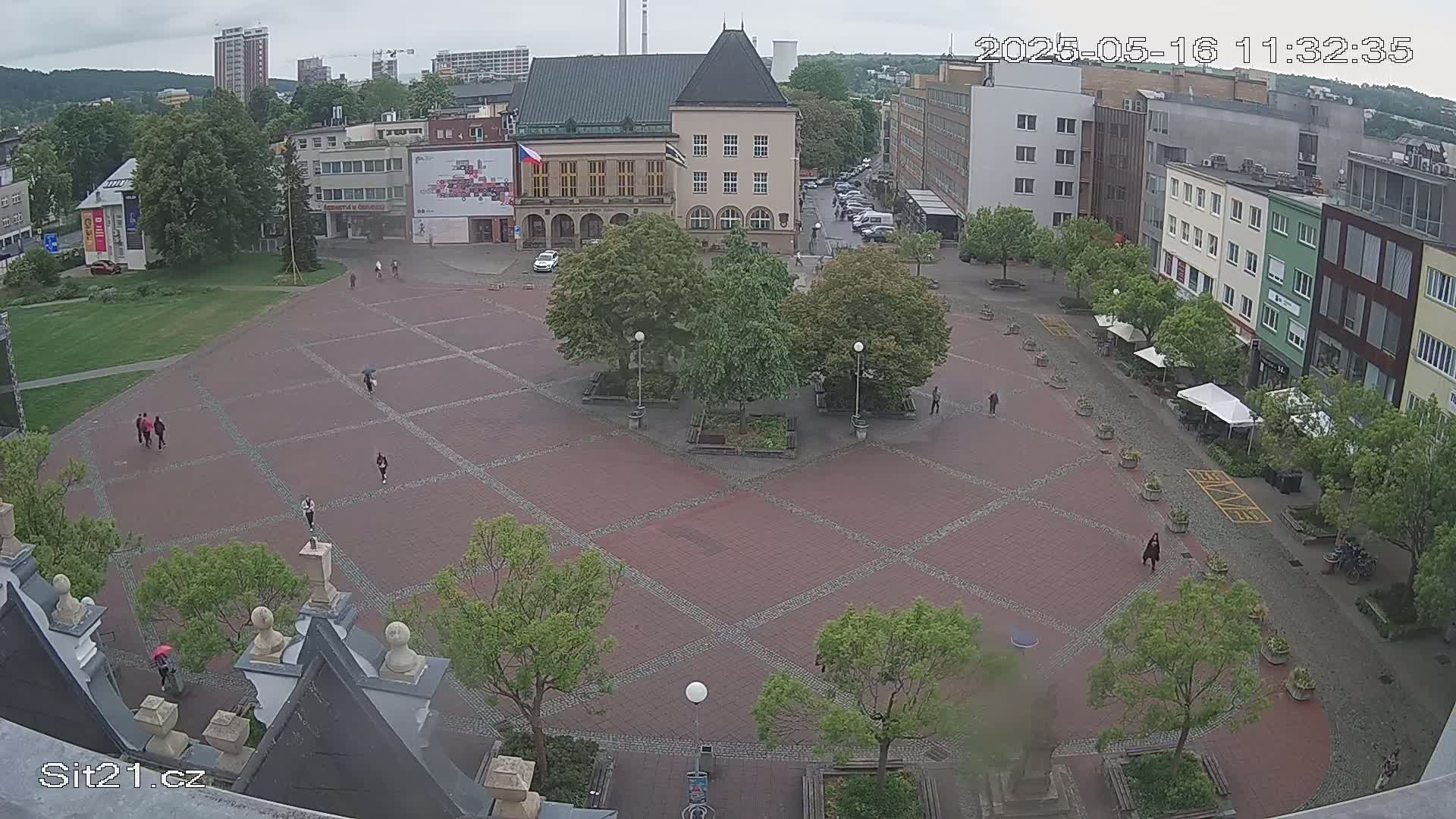 A brick town square with several people walking around, surrounded by buildings and trees under an overcast sky.