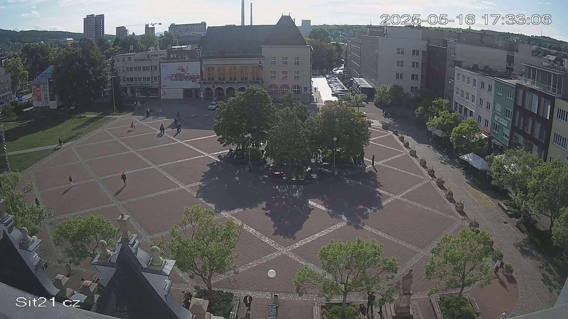 A paved town square with several buildings and trees surrounding it, under sunny conditions.