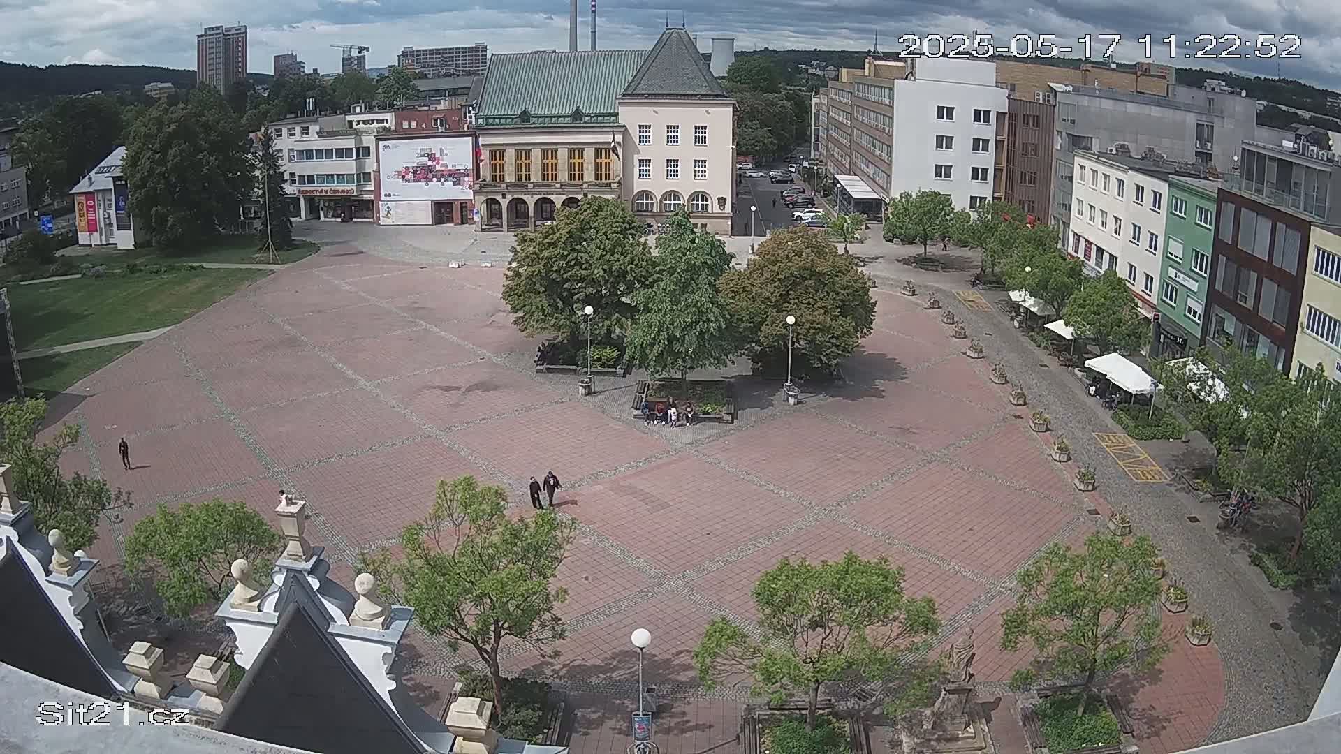 A mostly empty brick town square is surrounded by buildings under partly cloudy skies, with several people and some outdoor seating visible.