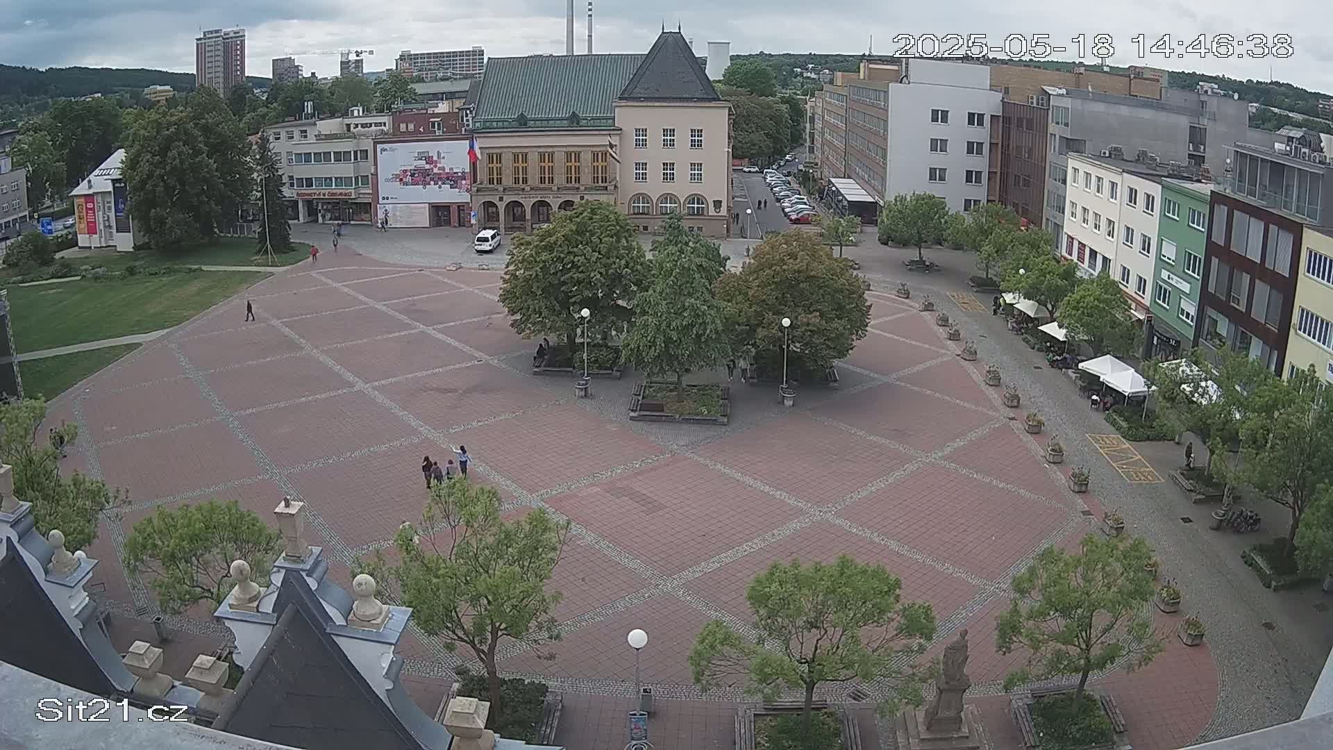 A brick town square with several buildings surrounding it, featuring green trees, benches, and a few people; the sky is overcast.