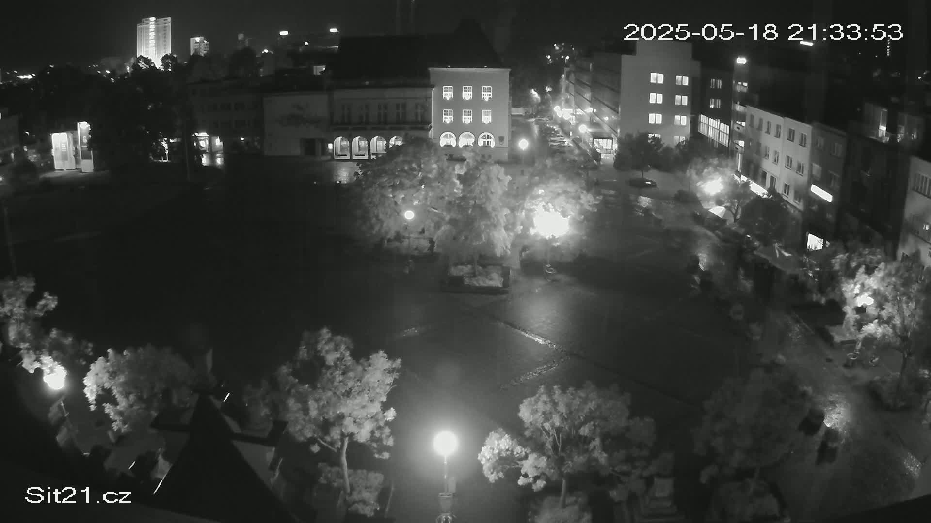 A nighttime, high-angle view of a town square showing wet pavement, buildings, and trees illuminated by streetlights under a dark sky.