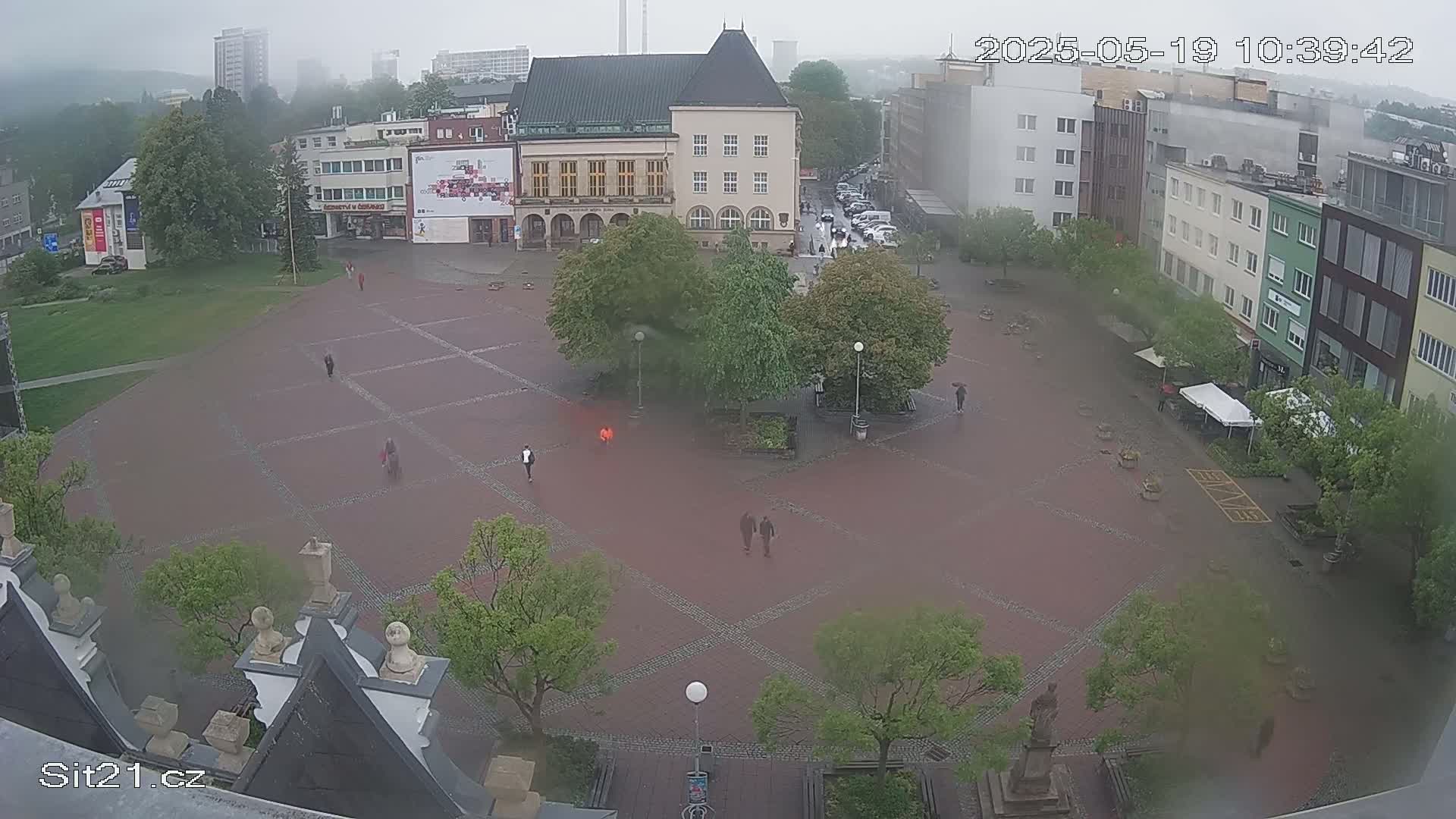 A rain-swept town square is seen from an elevated viewpoint, showing several buildings, trees, and a few pedestrians.